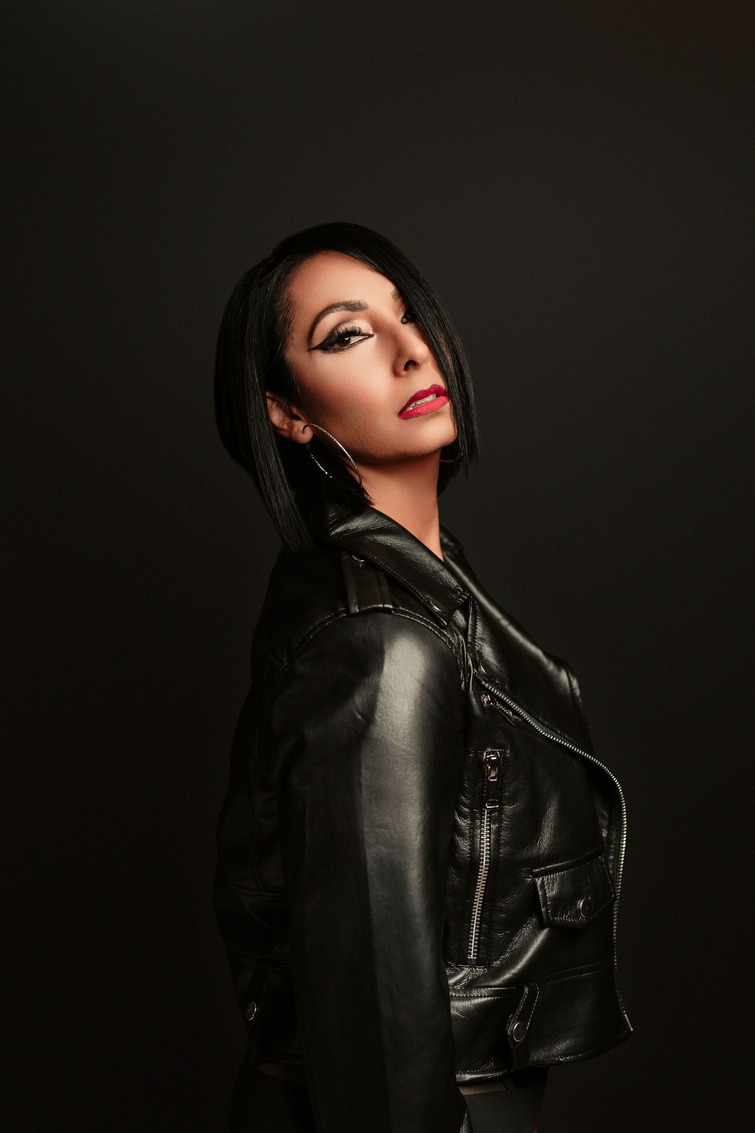 Dramatic fashion portrait in dark lighting shows model wearing black leather jacket against shadowy background.