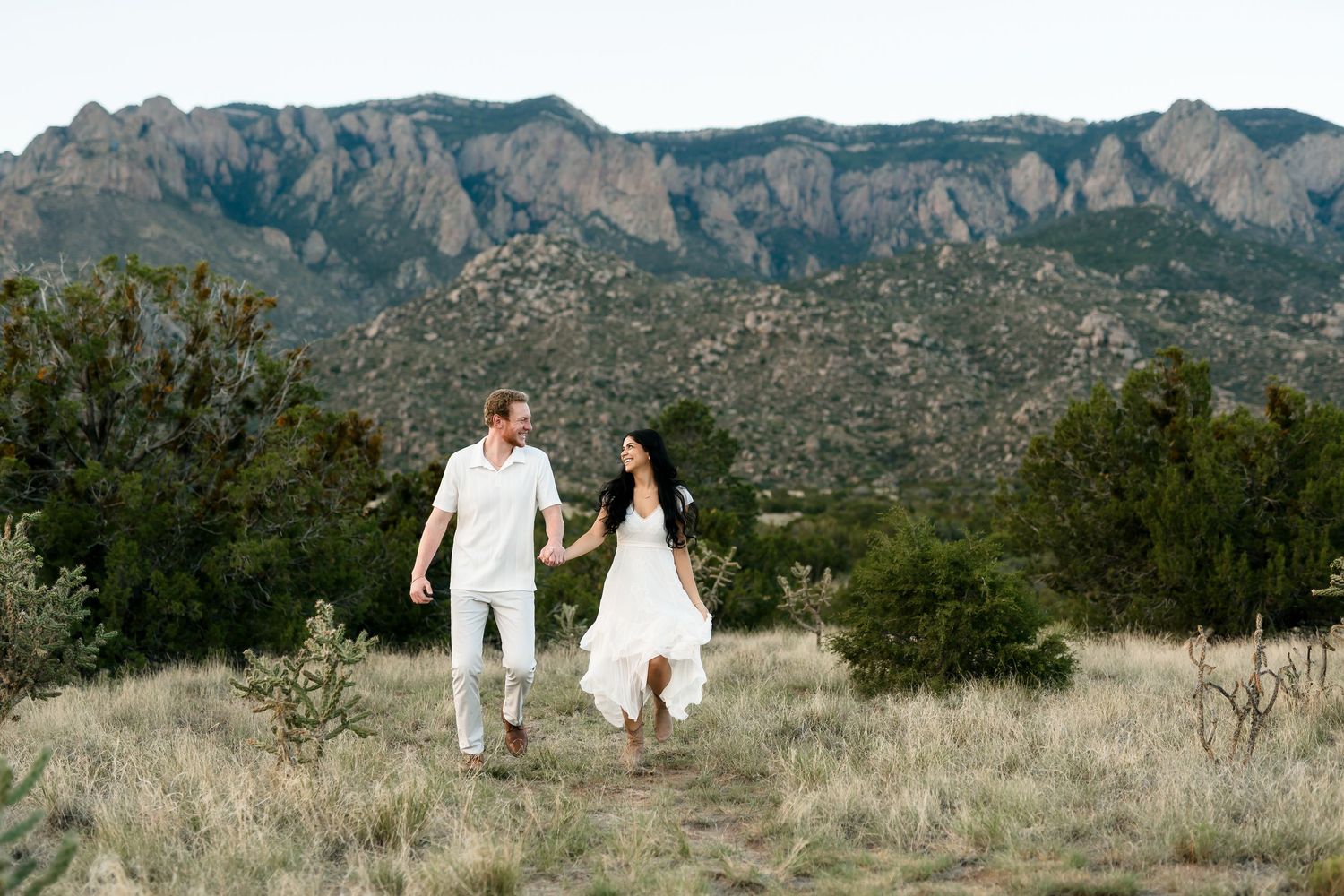Couple in white outfits holding hands walking through desert landscape with Sandia Mountains in background.