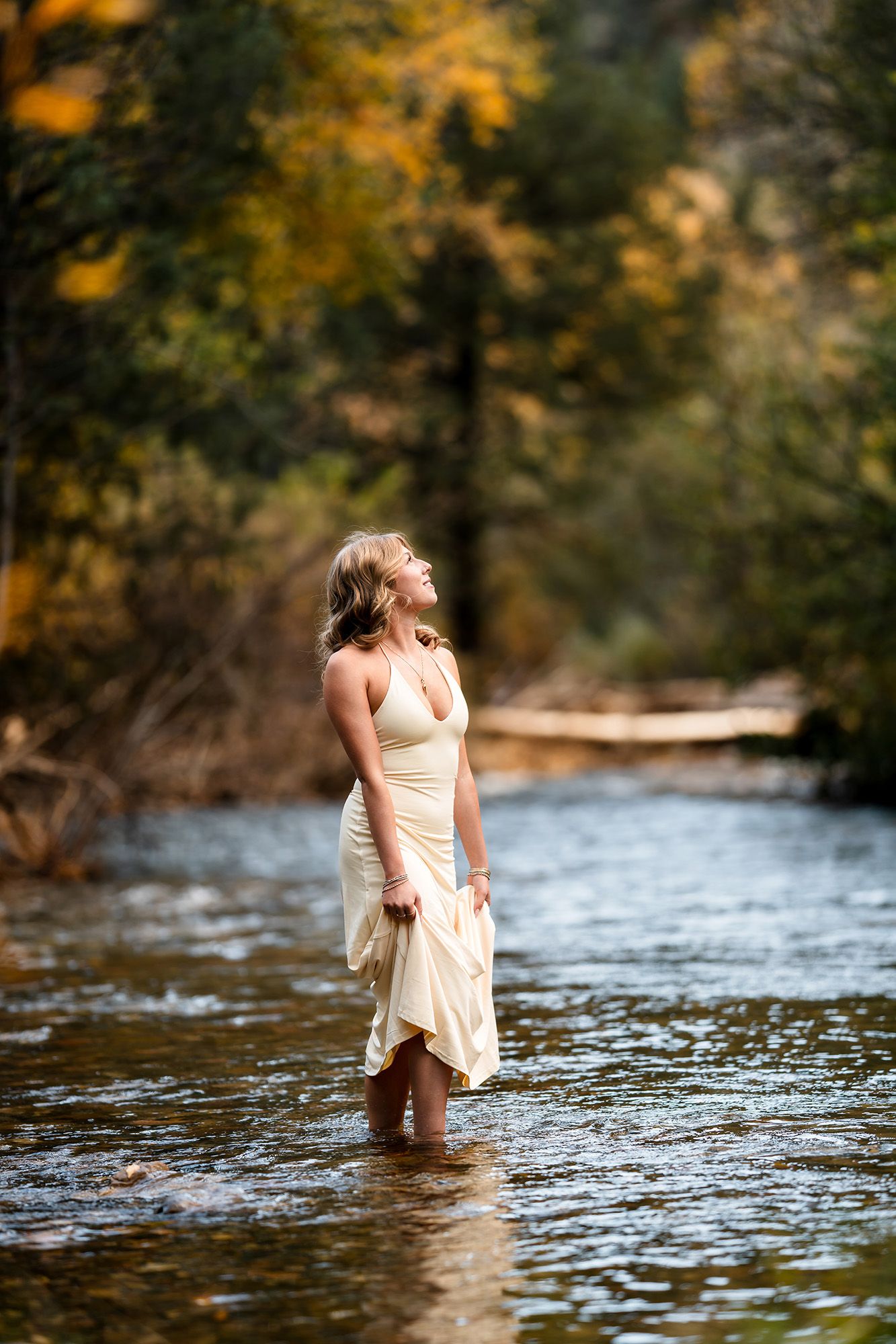 Woman in a flowing cream dress wading in a shallow river, gazing upward, with golden autumn foliage glowing in the forest behind her.