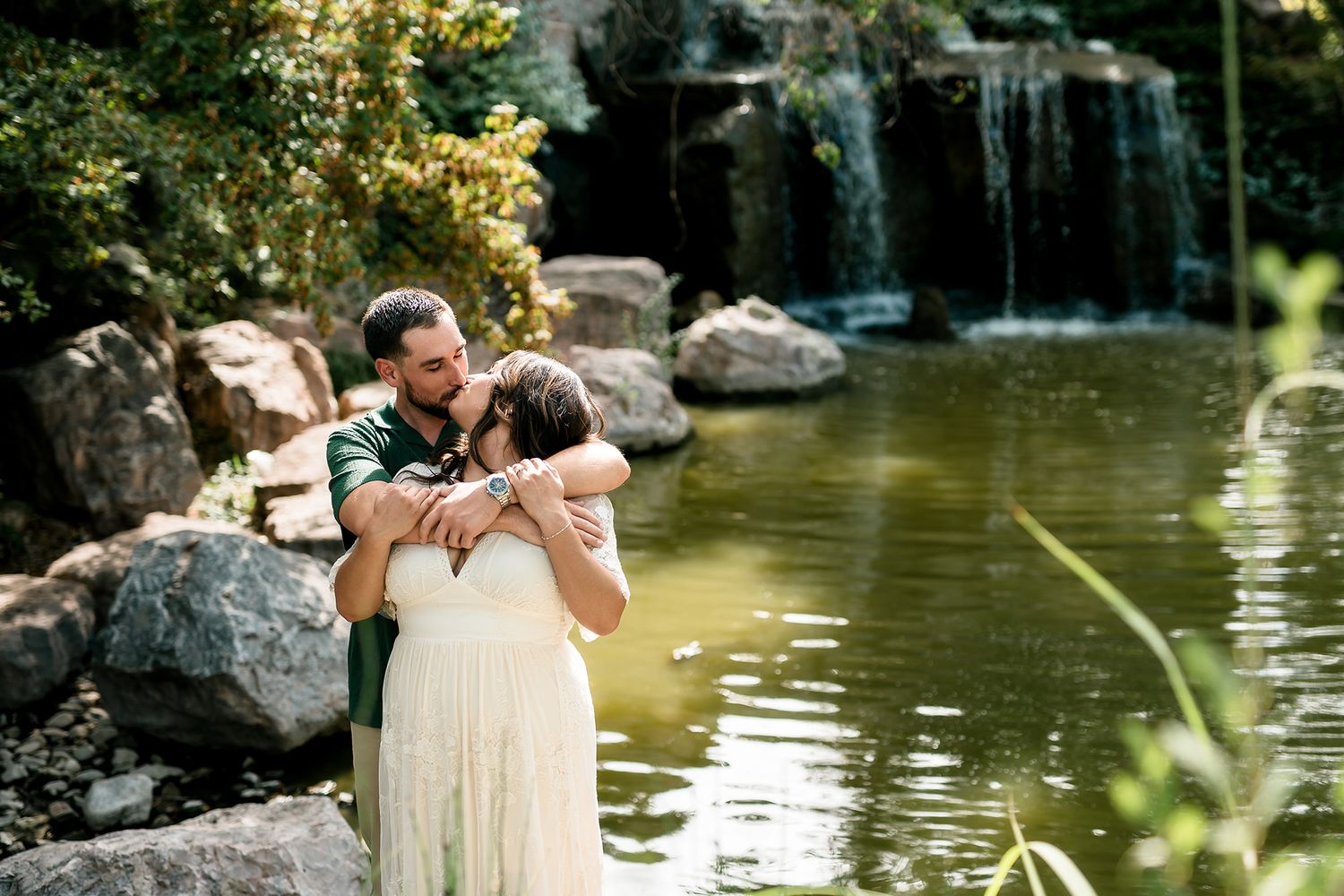 Couple embracing romantically by a serene garden waterfall pond, surrounded by lush greenery and rocks.