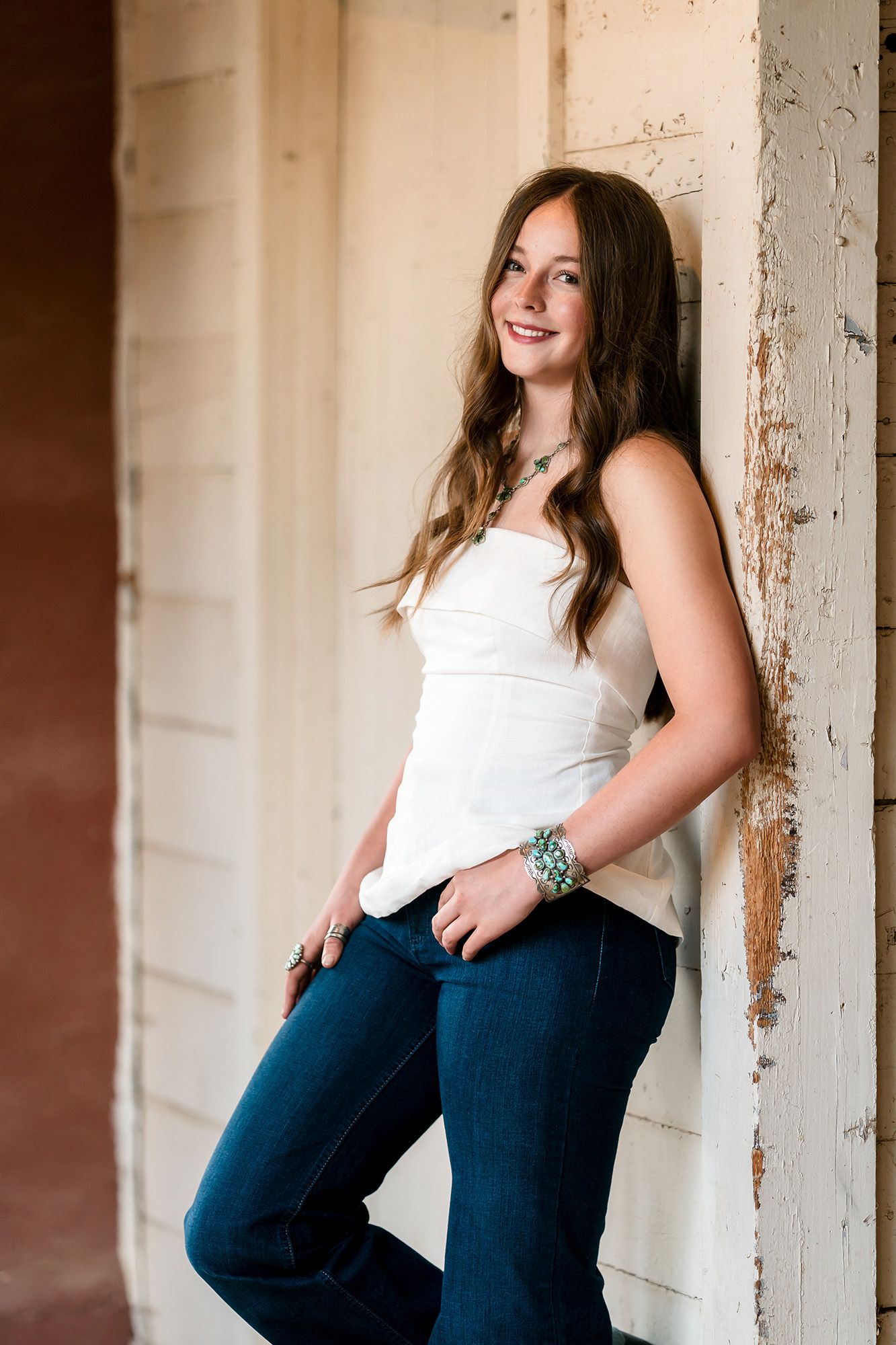 Young woman in white tank top and jeans leaning against a rustic white wall, smiling confidently.