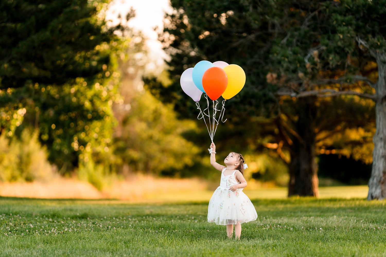 Young girl in white dress holding colorful balloons in a sunny green park with tall trees in the background.