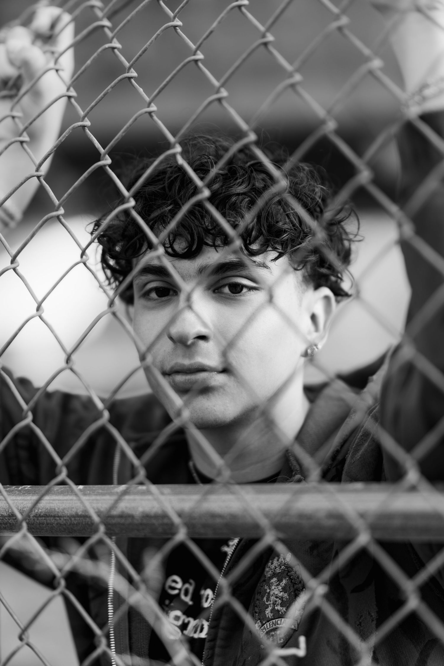 Monochrome portrait through chain link fence showing curly hair and urban style.