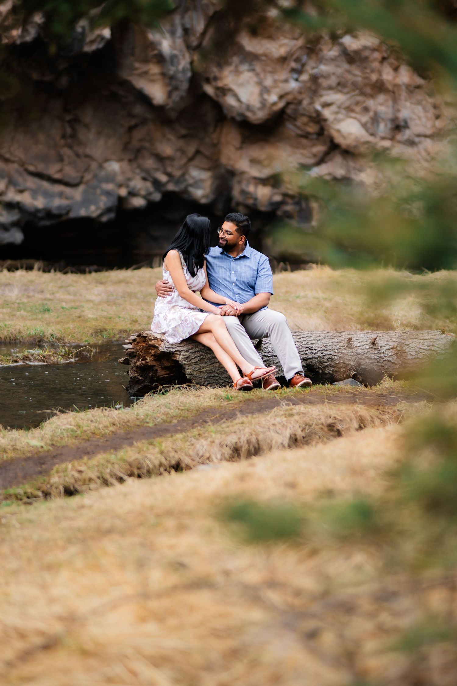 A couple sits together on a fallen log by a stream with rocky cliffs in the background.