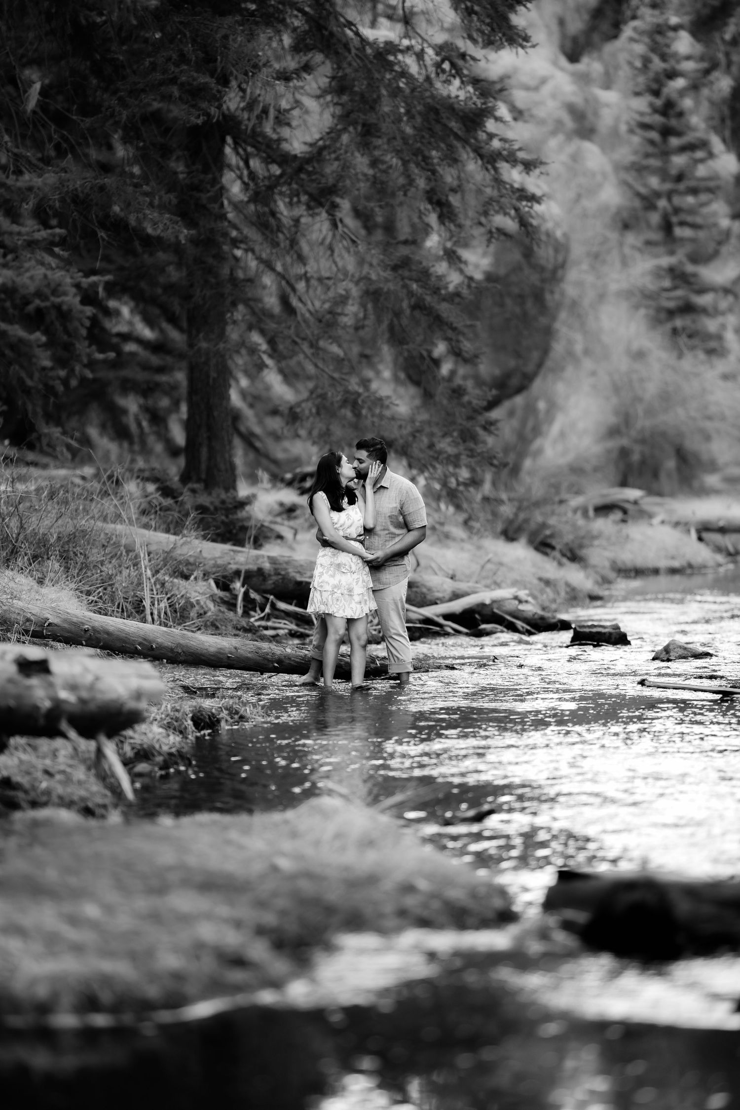 A couple sharing a tender moment by a stream in a forest setting captured in black and white.