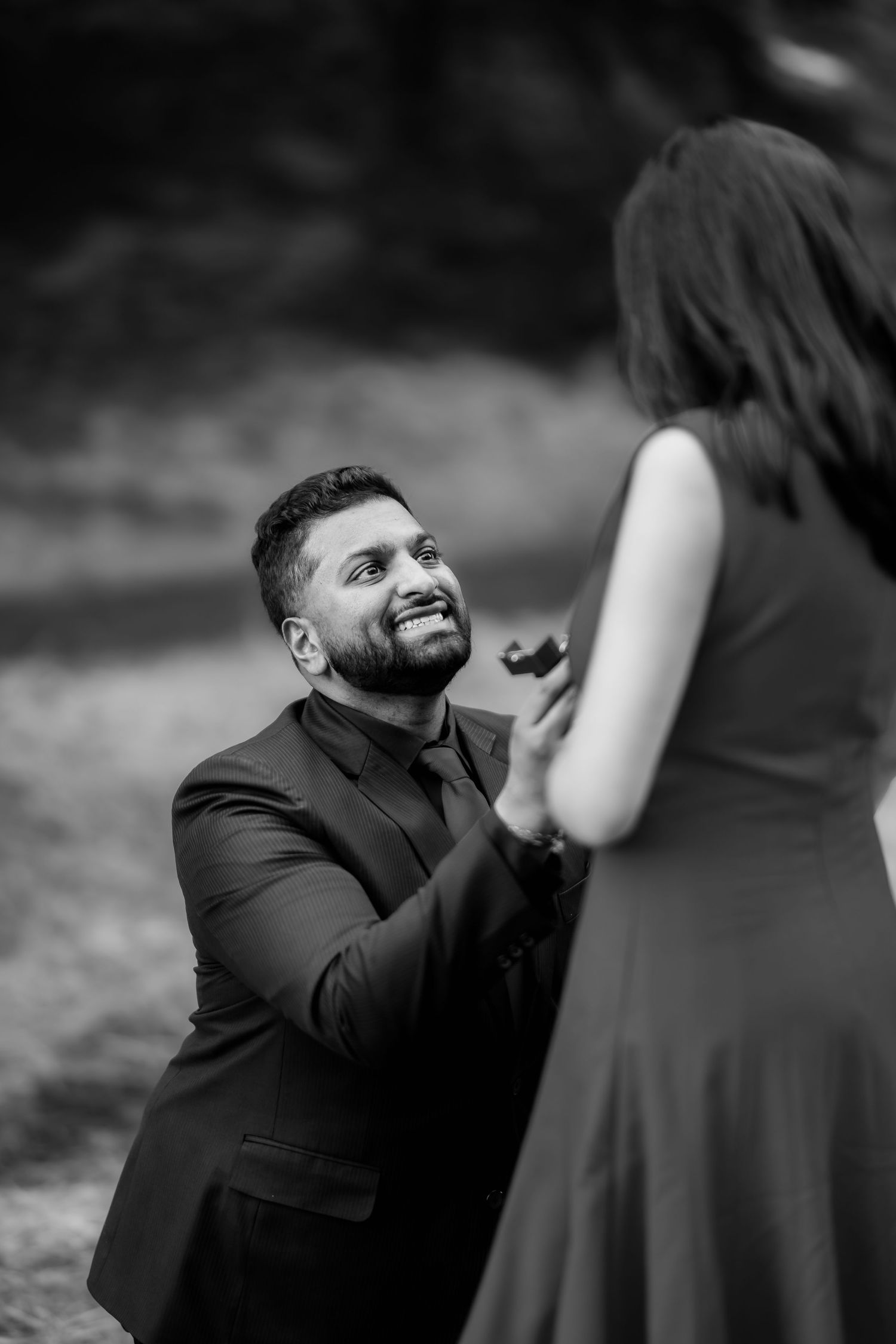 A romantic black and white proposal moment captured outdoors with a kneeling figure presenting a ring.