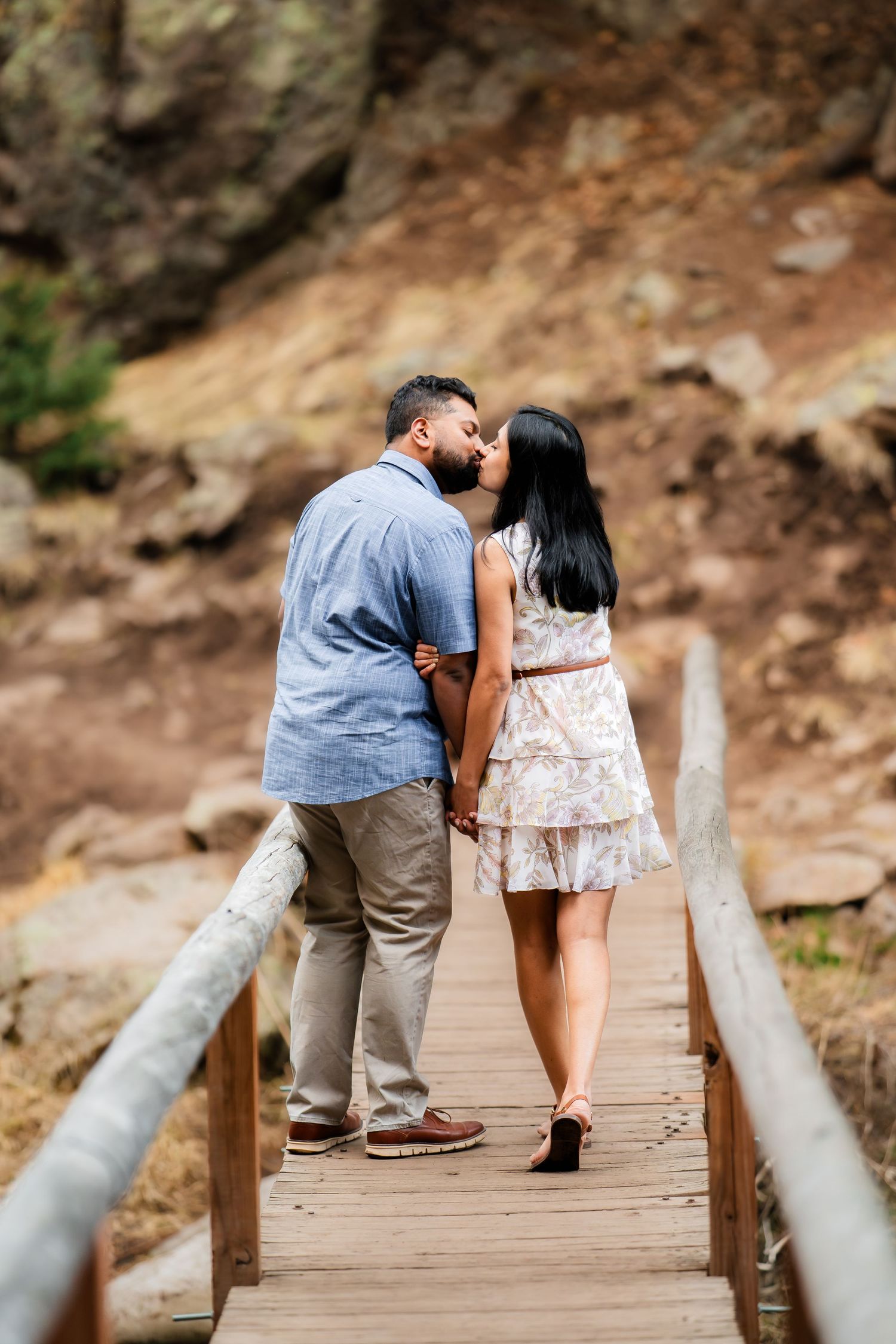 Intimate couple shares moment on wooden bridge with rocky mountain backdrop.