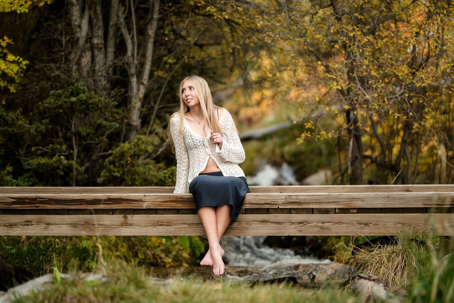 Blonde woman in cream sweater and skirt sits barefoot on wooden bridge surrounded by autumn foliage.