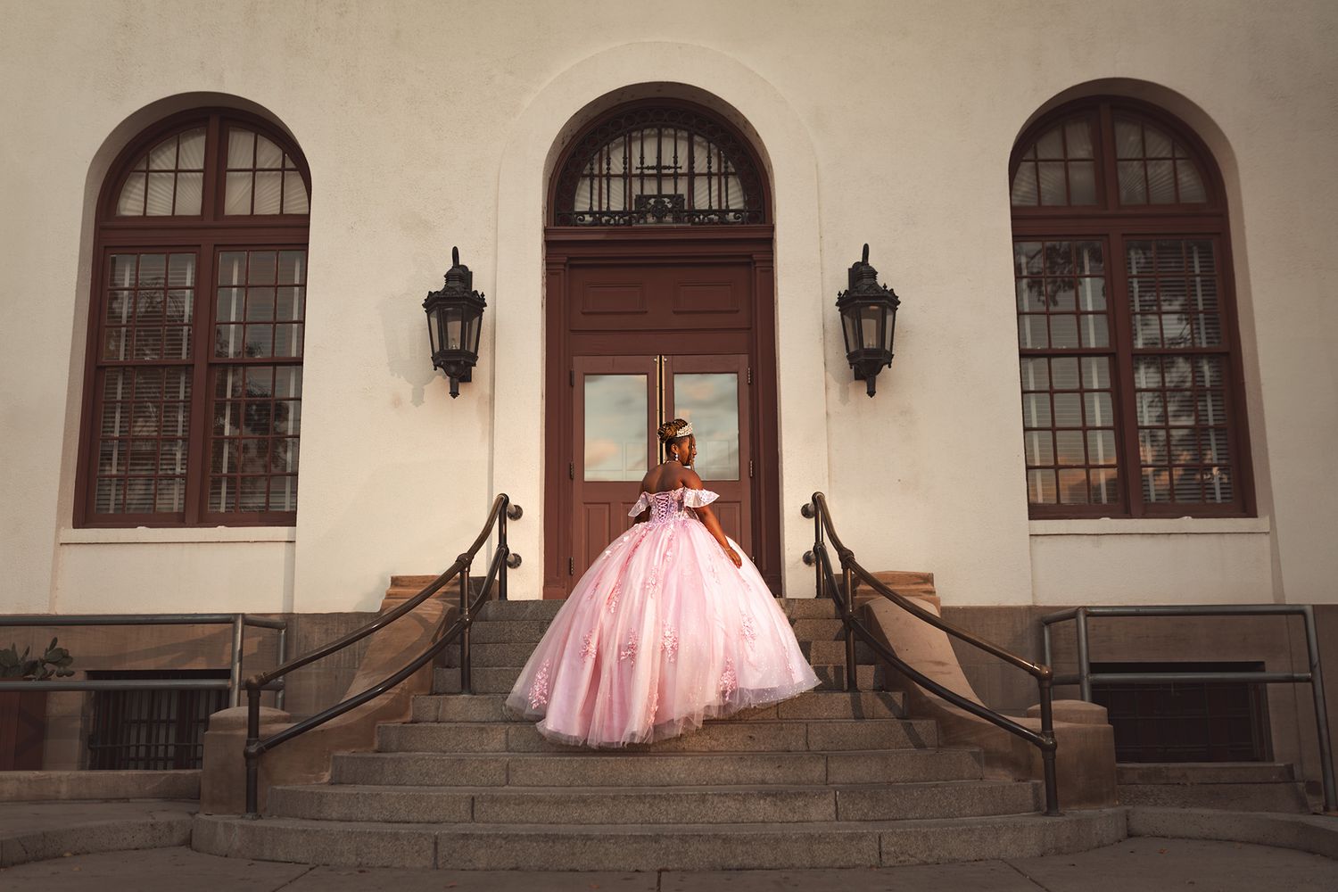 A person in a voluminous pink ballgown stands elegantly on stone steps in front of an arched doorway with vintage lanterns.