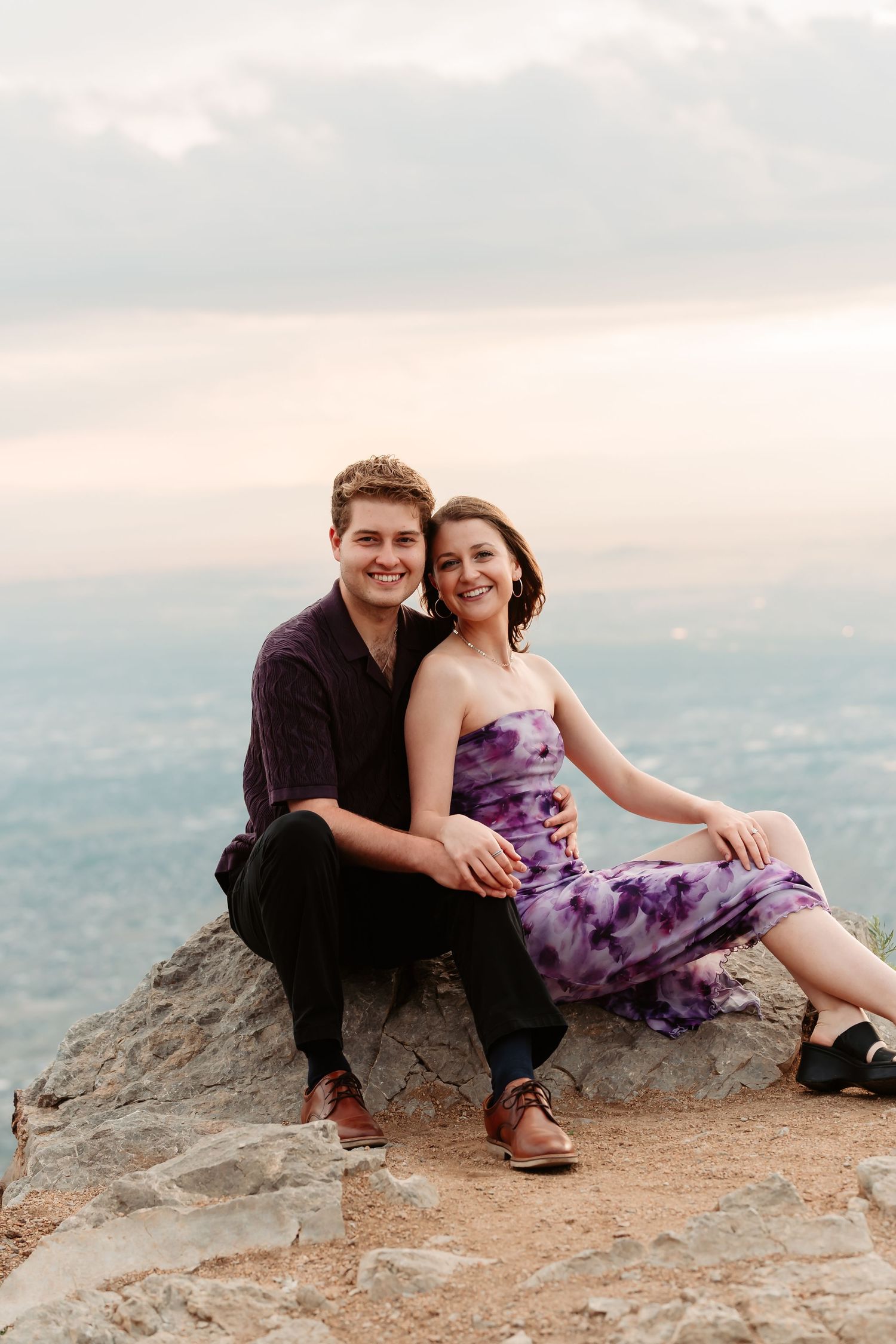 Couple sits together on mountain ledge overlooking vast valley landscape.