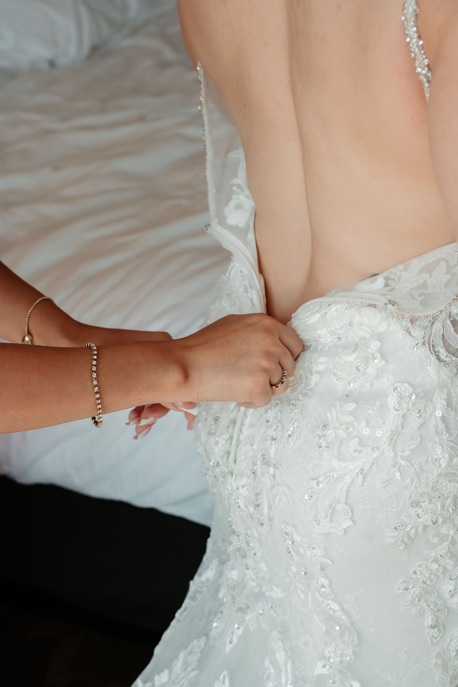 Close up of someone helping button up an intricate white lace wedding dress against light bedding.