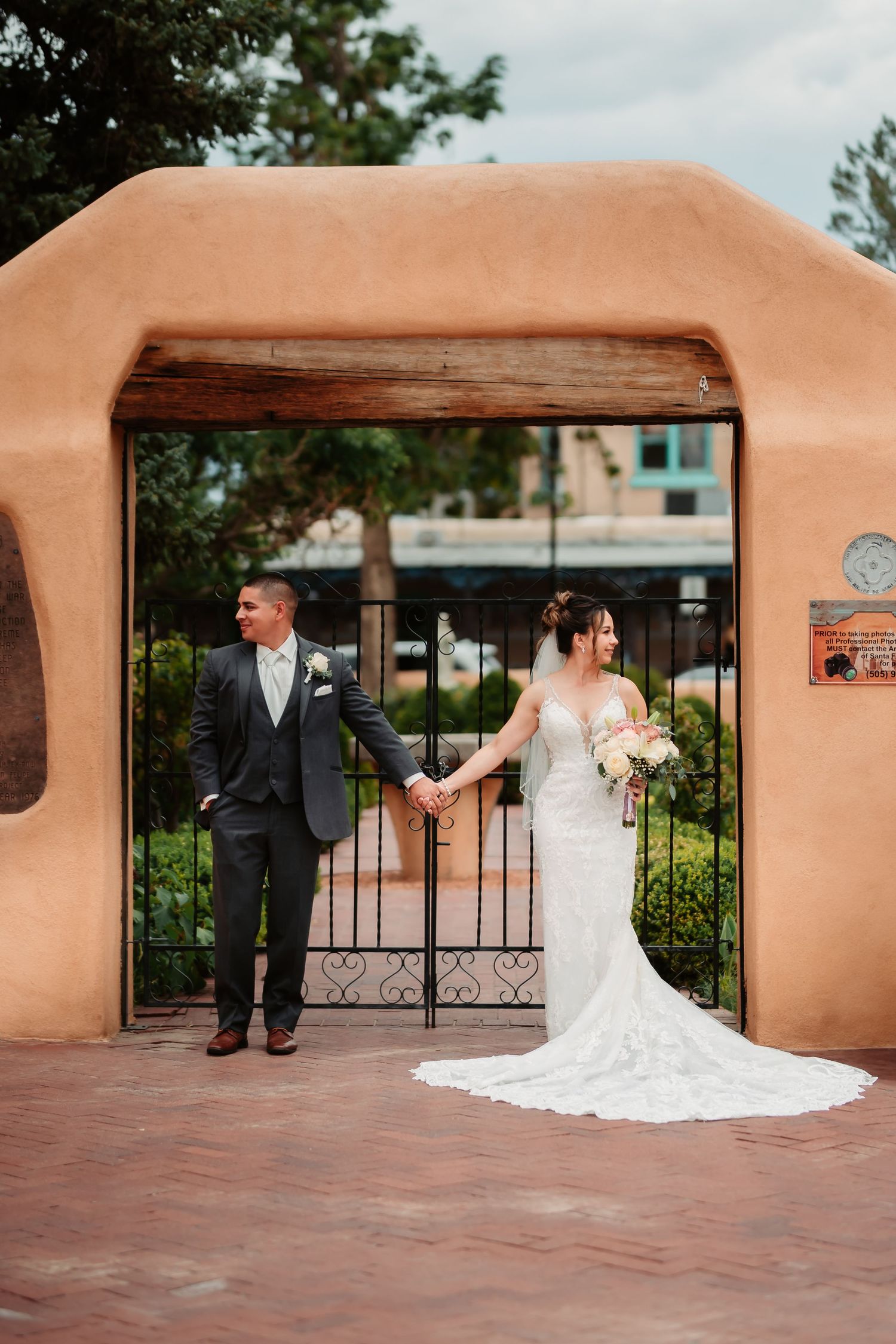 A bride and groom pose against a southwestern style adobe wall and iron gate.