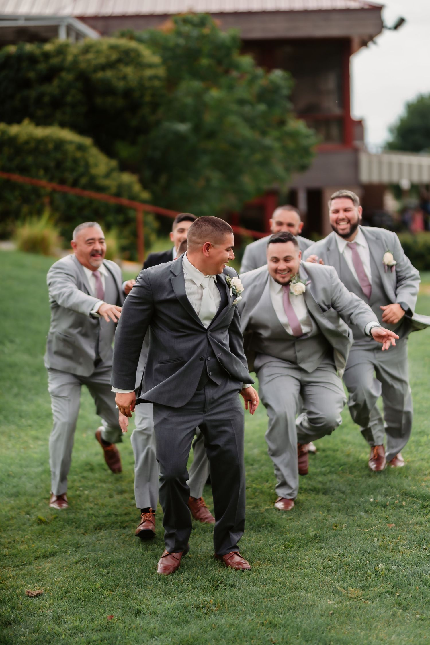 Wedding party members in gray suits charge forward dramatically on grass during fun photo session.