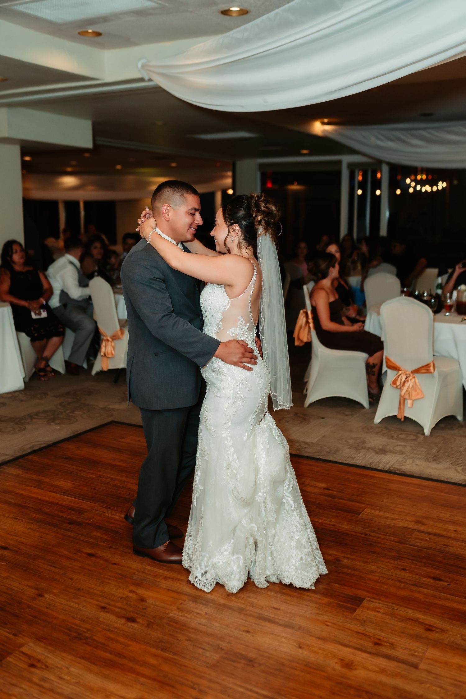 A couple shares their first dance as newlyweds on a wooden floor under elegant white draping at their wedding reception.