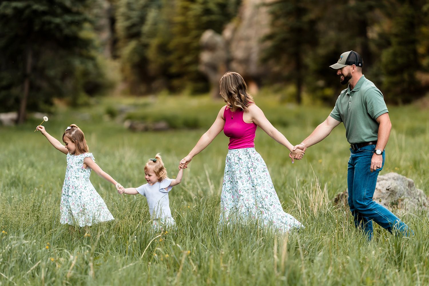 Family of four holding hands walking through green meadow with trees in background.