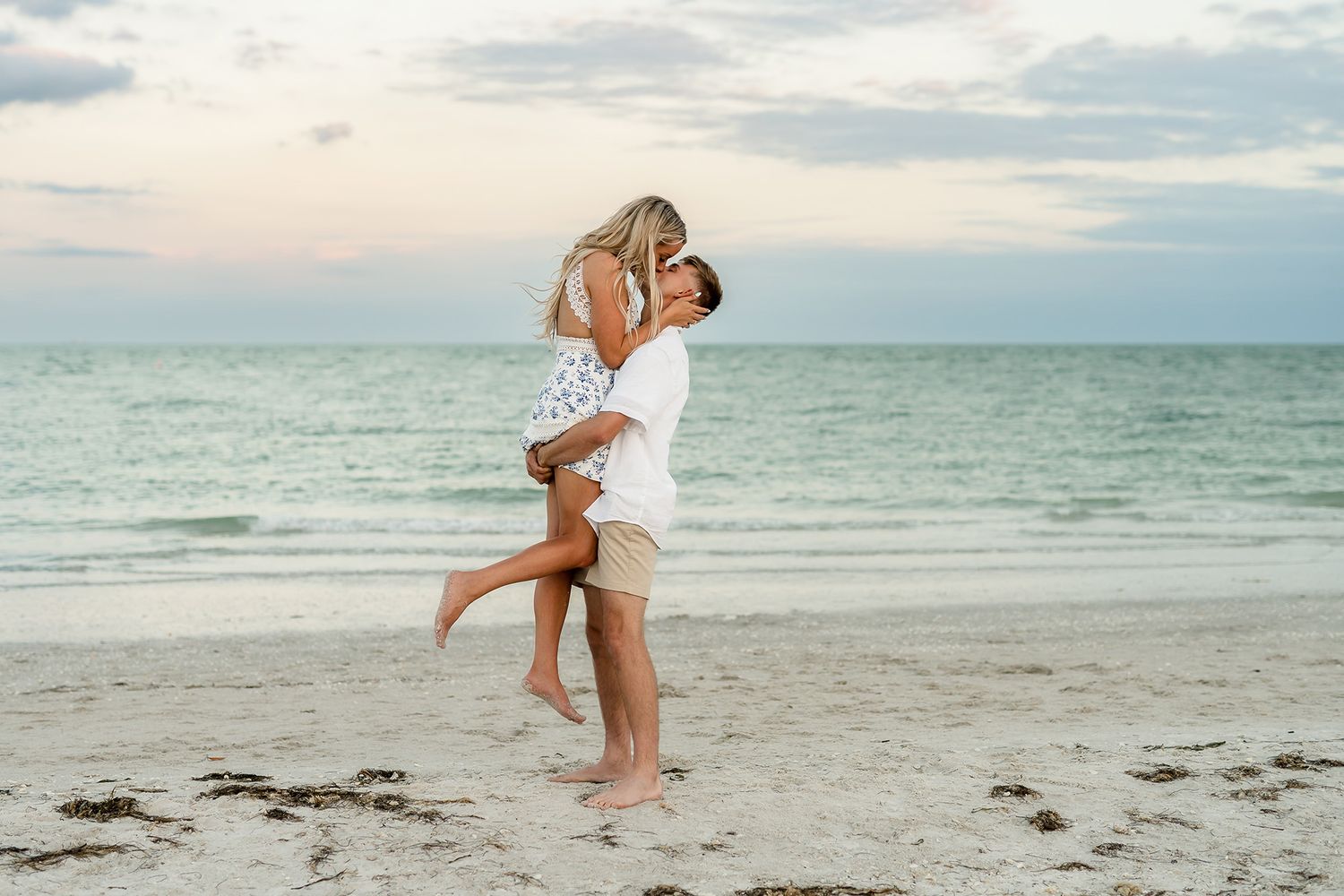Couple embracing on a sandy beach at sunset, man lifting woman in romantic pose with ocean waves in background.