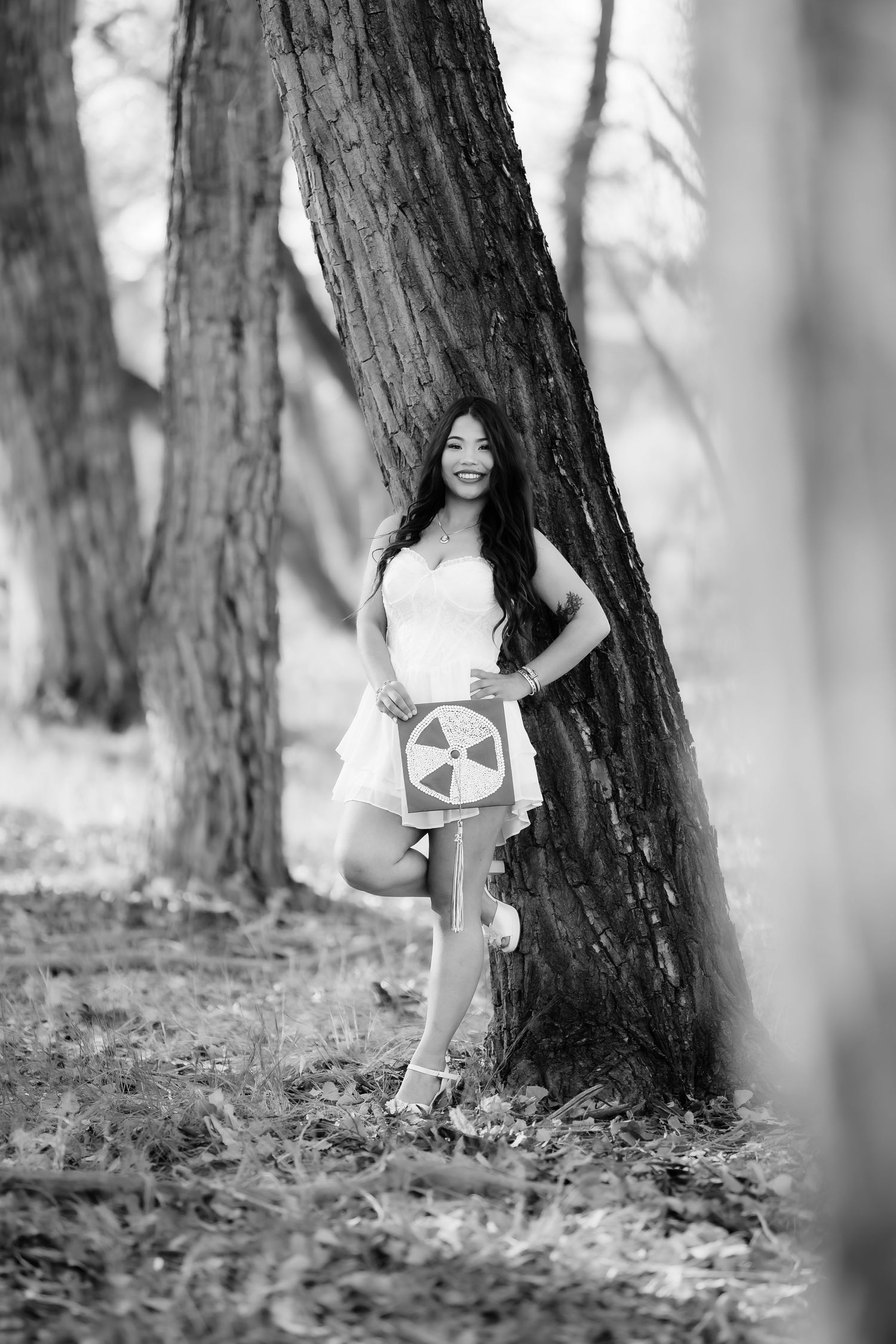Black and white portrait of person in white dress posing between trees in forest setting.