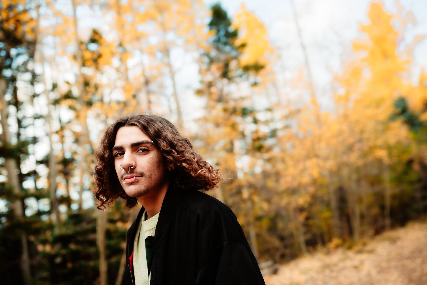 Person with curly hair poses outdoors against autumn foliage and evergreen trees in natural lighting.