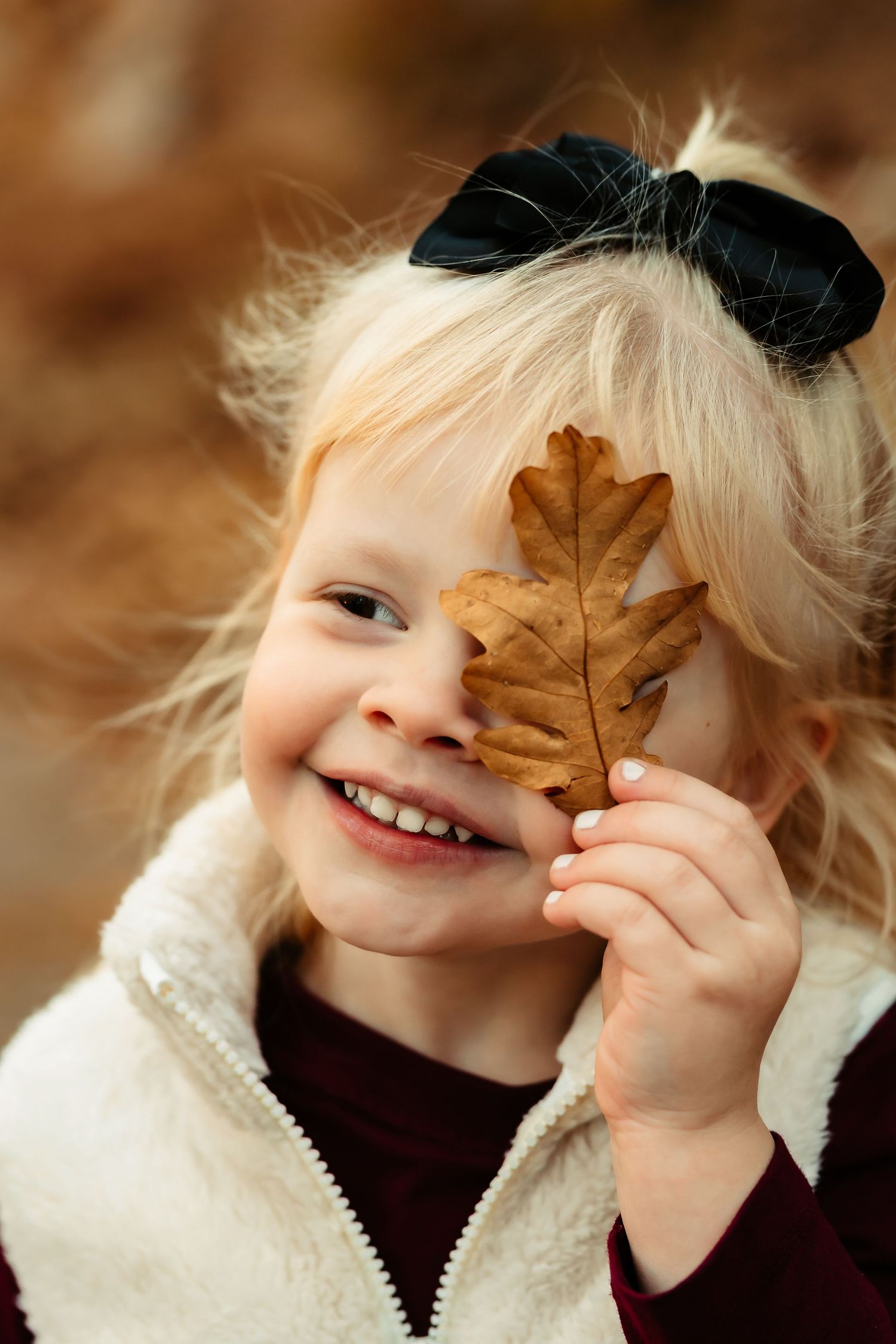 A child playfully holds an autumn leaf over their face while wearing a white sweater against a warm brown background.