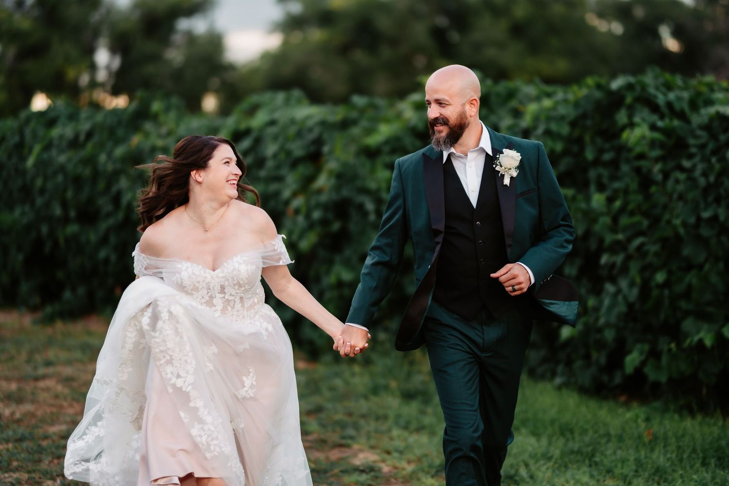 A couple in wedding attire runs joyfully through a garden setting during their outdoor celebration.