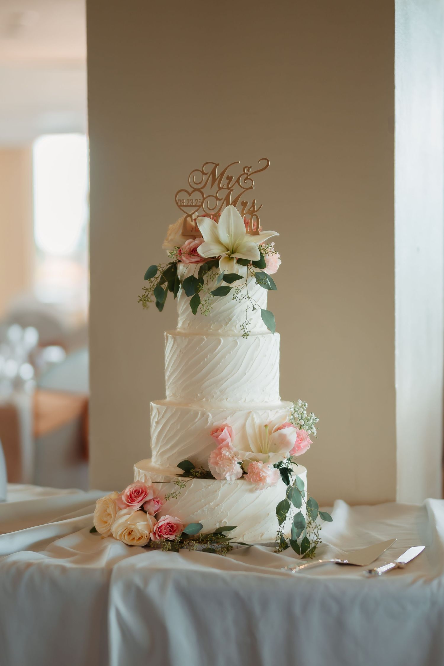 Elegant white tiered wedding cake decorated with pink roses and greenery displayed on a white tablecloth.