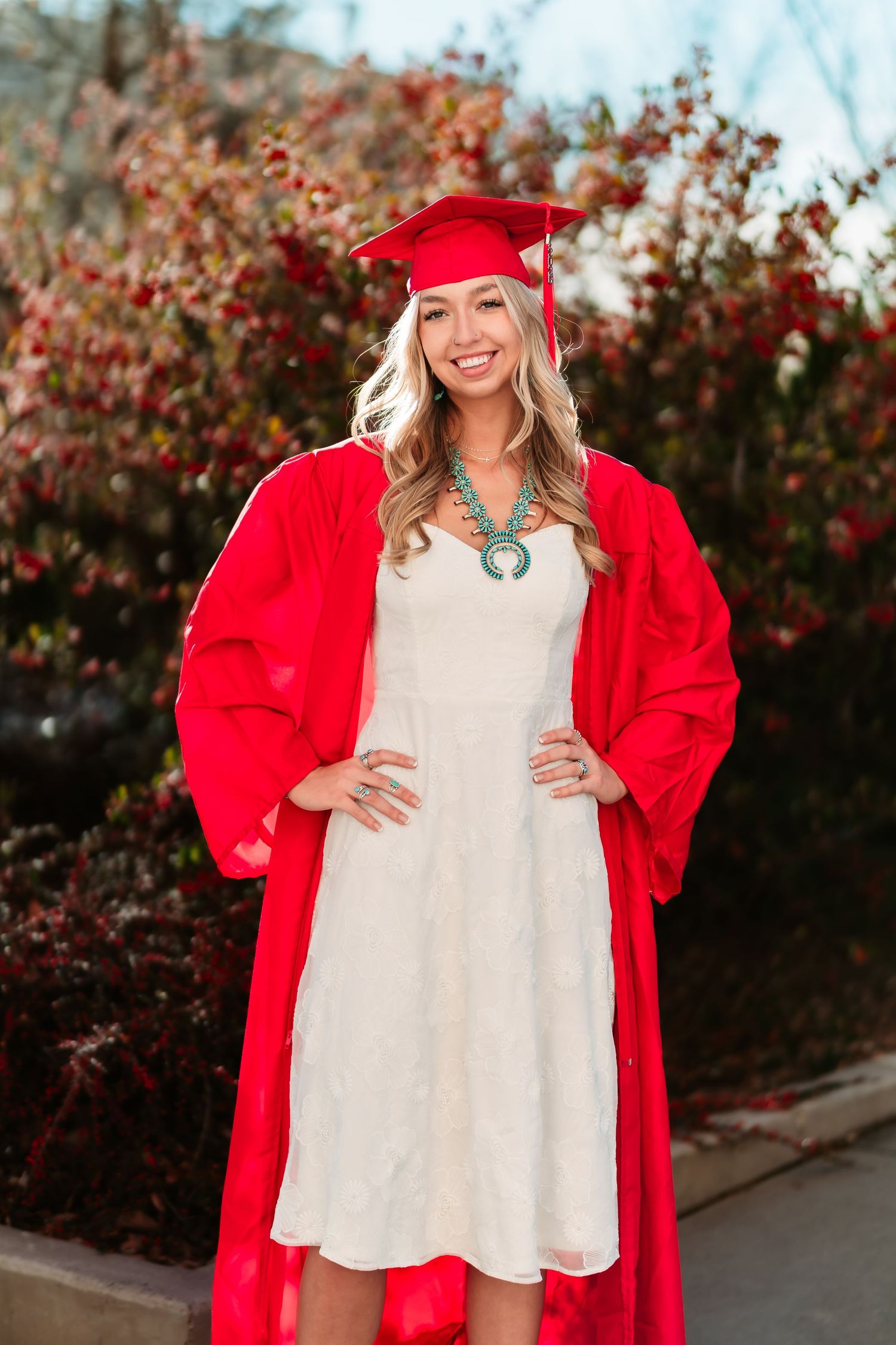 Graduate in bright red cap and gown poses joyfully against a backdrop of autumn foliage.