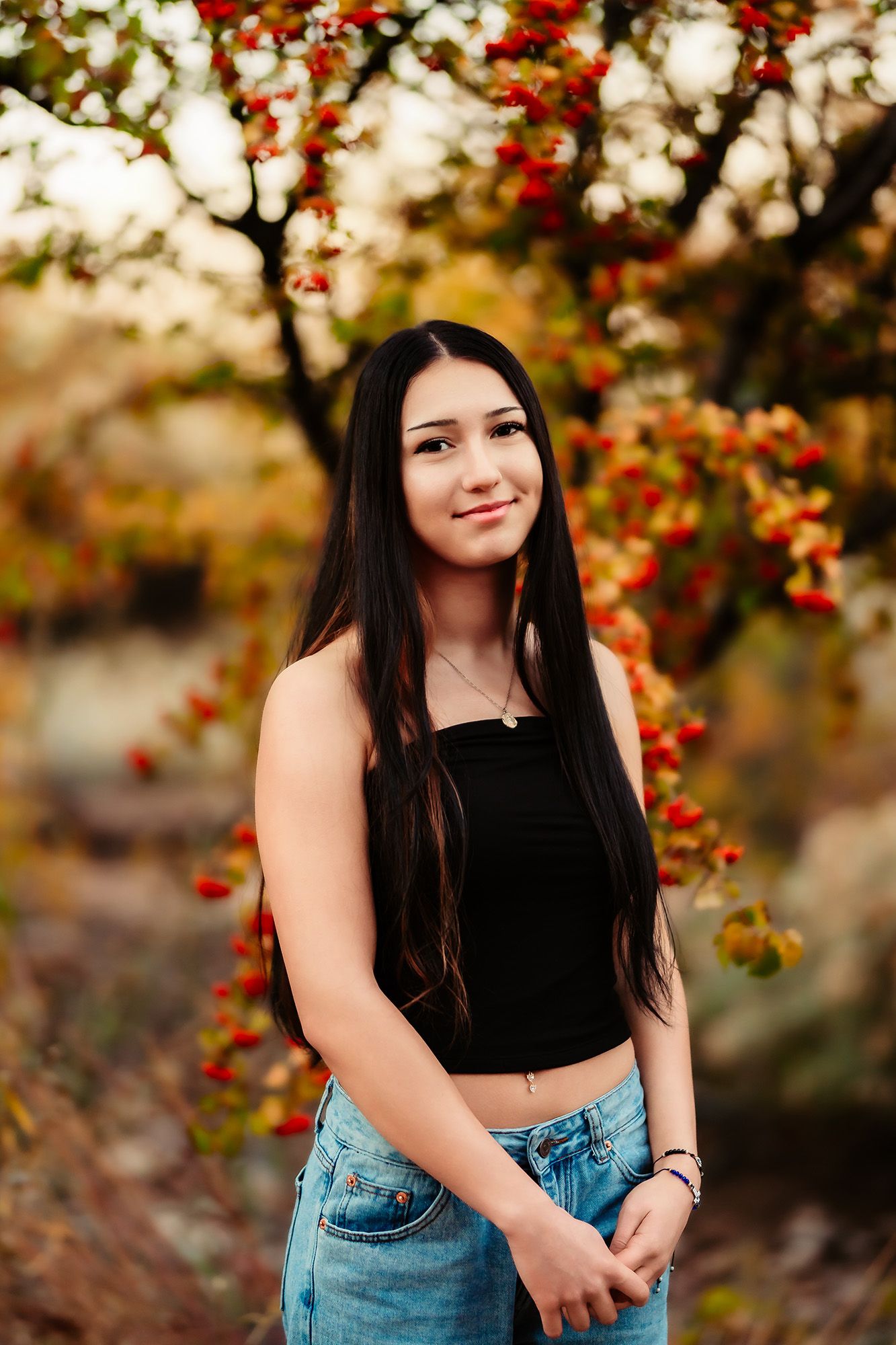 Portrait of someone in a black crop top and denim shorts posing outdoors with autumn foliage in the background.