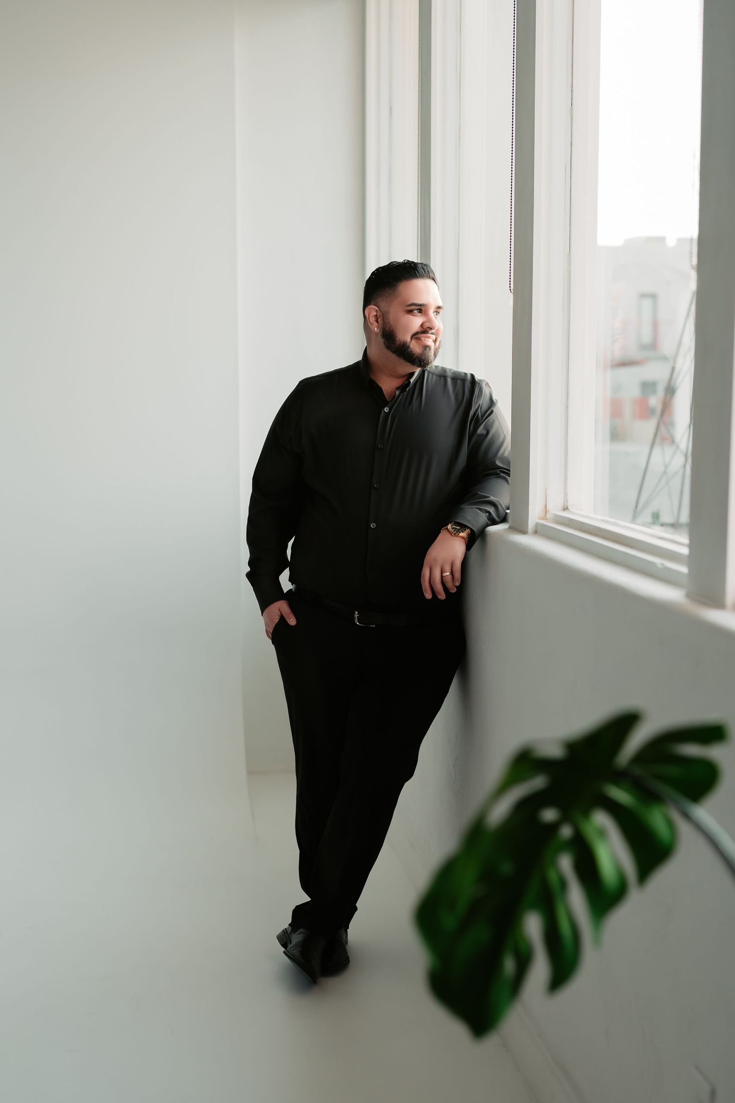 A person in all black attire leans against a bright white wall near a window with a monstera plant in the foreground.
