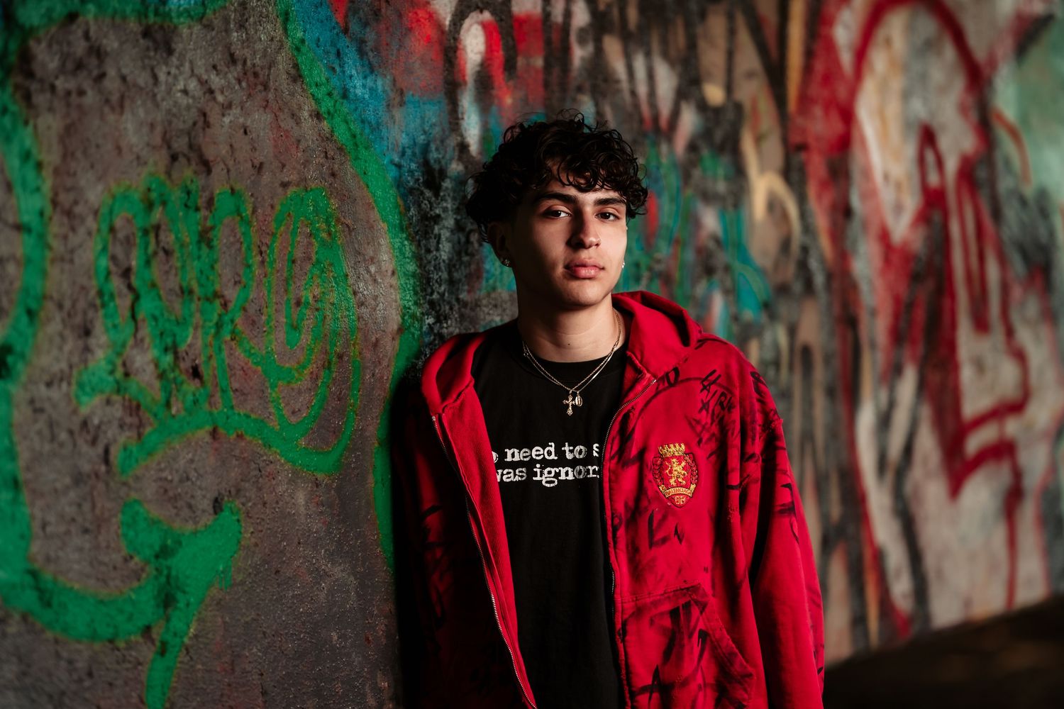 A person in a red hoodie stands against a graffiti-covered concrete wall with dramatic lighting.