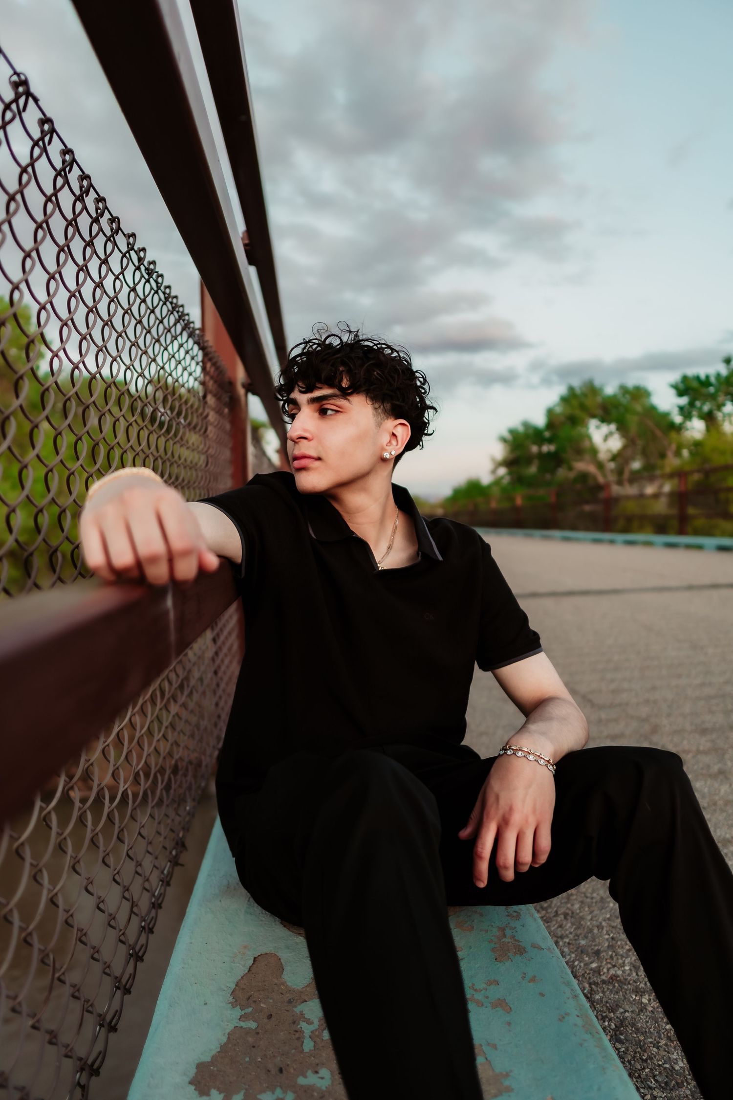 Artful portrait of someone in black clothing sitting against chain-link fence during cloudy sunset.