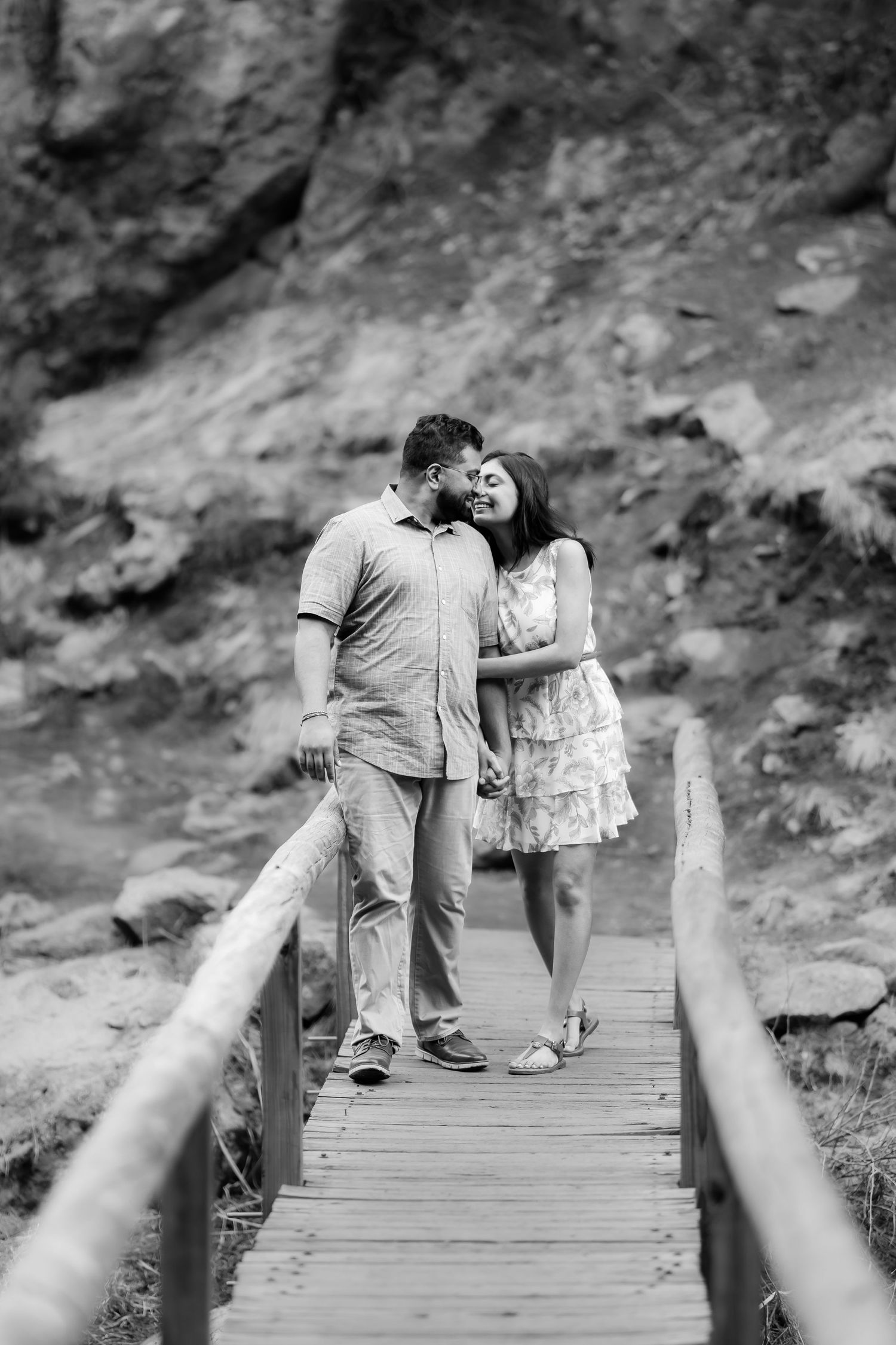 A romantic couple shares an intimate moment while standing on a wooden bridge against rocky cliffs.