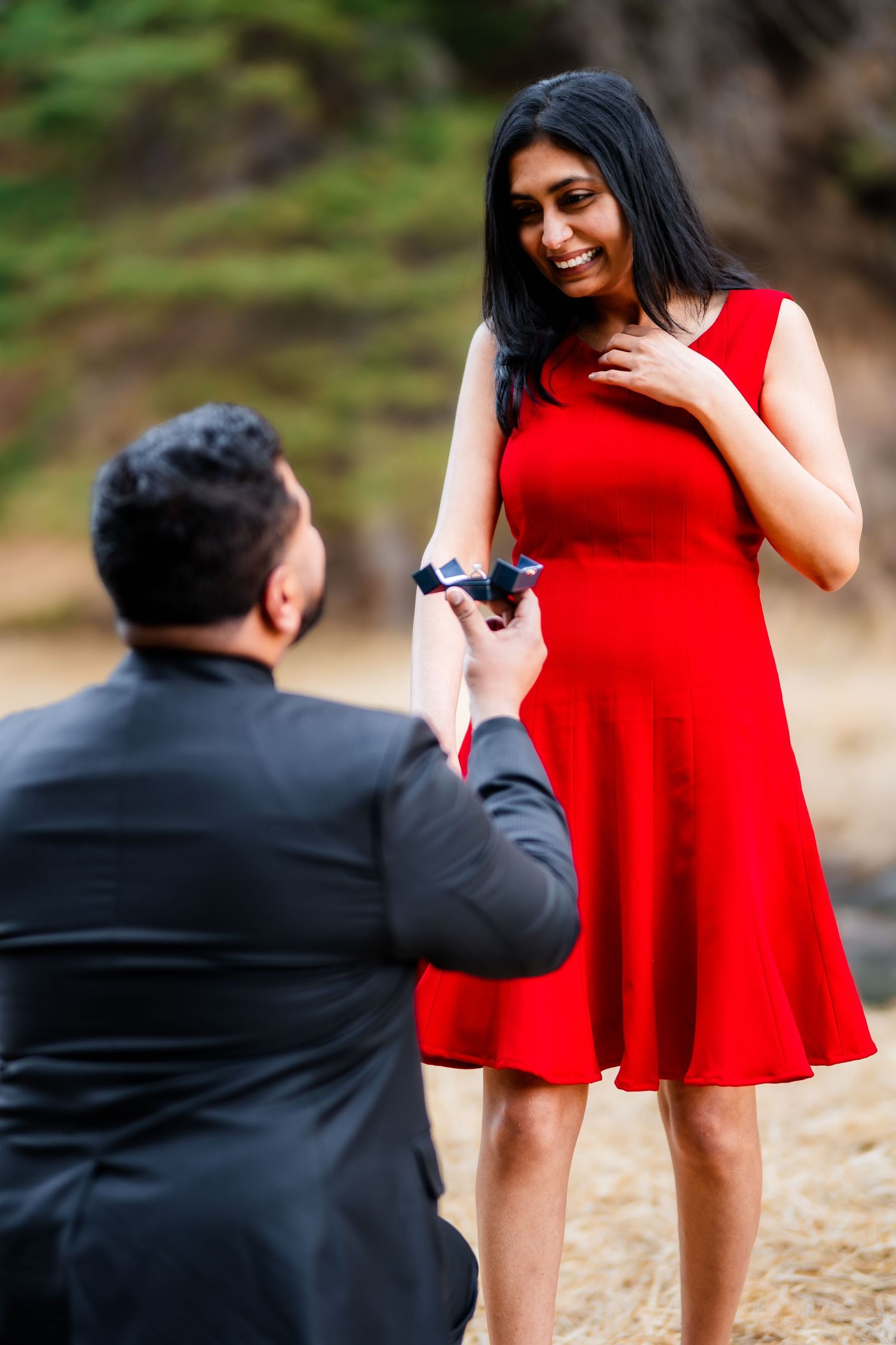 A proposal moment captured in nature as a person in black kneels before another wearing a red dress.