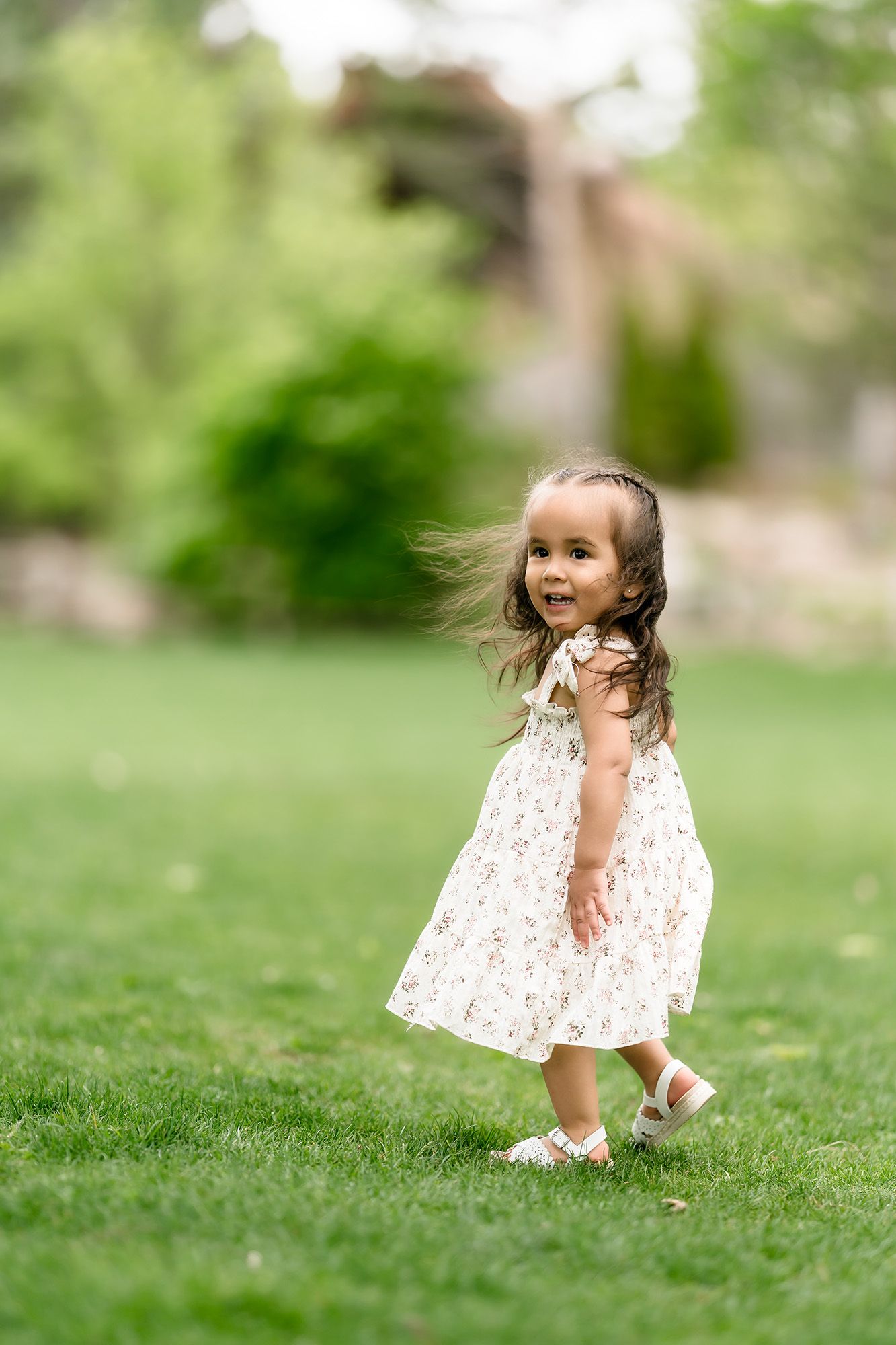 Toddler girl in floral dress playing on green grass in a park on a sunny day.