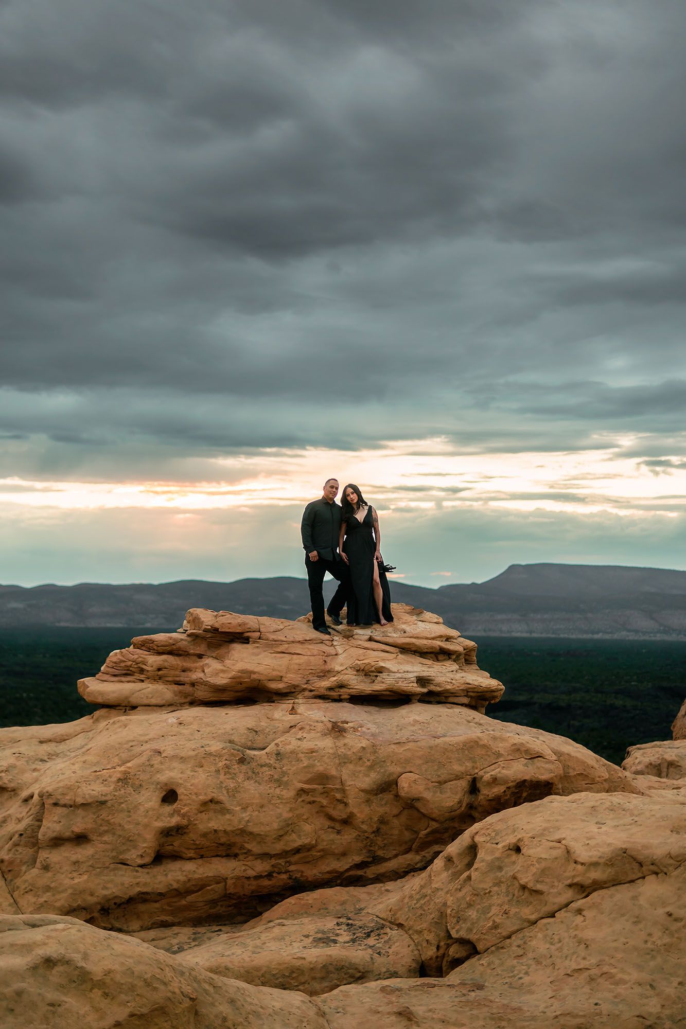Couple dressed in black standing together on sandstone rock formation under dramatic cloudy sky.