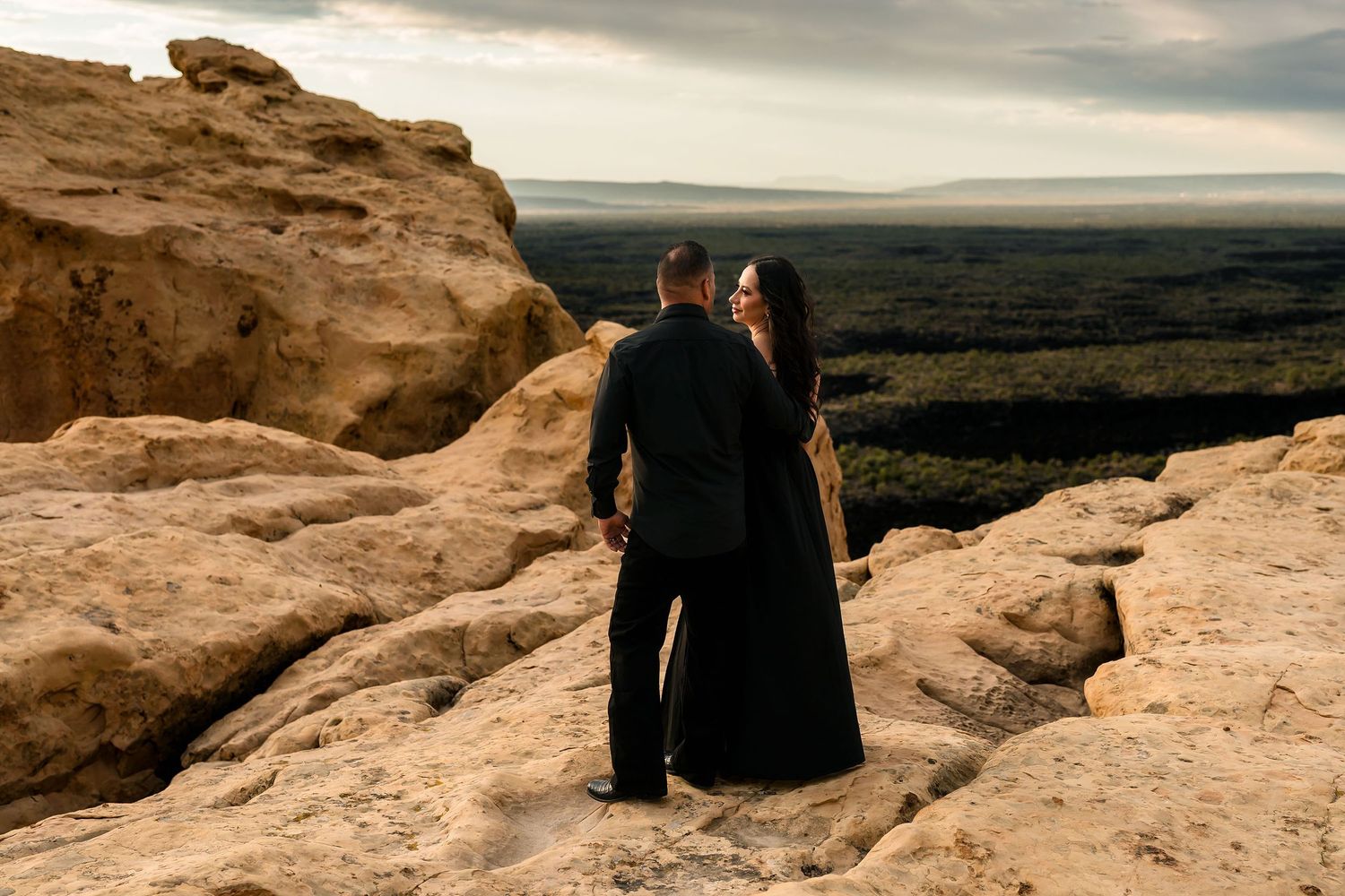 Couple in black standing on sandstone cliff edge overlooking a vast green desert landscape below.