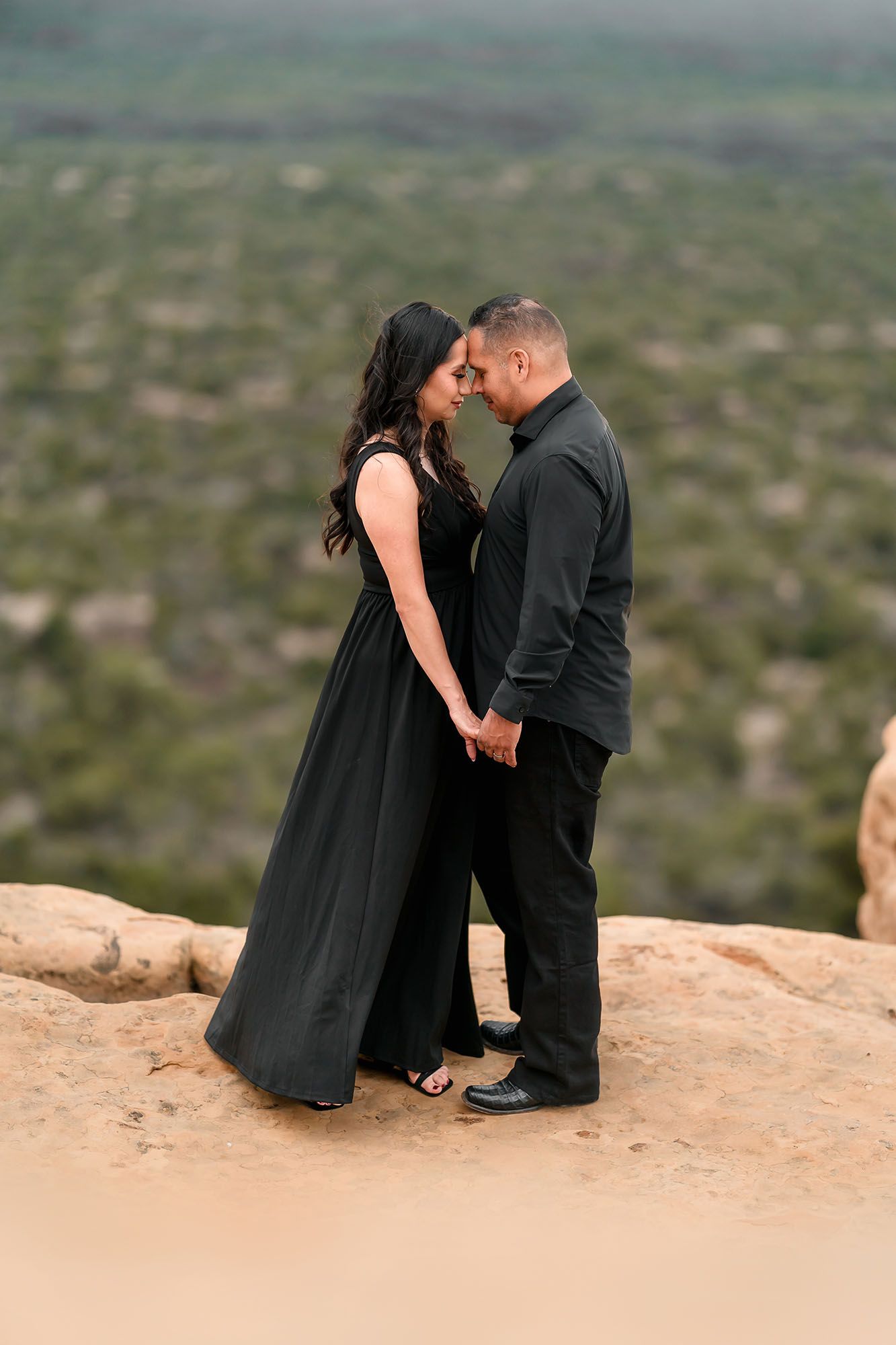 Couple in black formal wear holding hands on rocky overlook with green valley below.