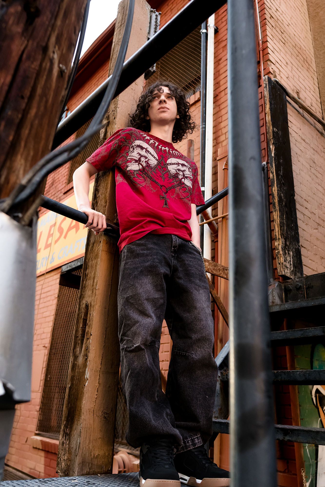 Young man in red graphic tee and black jeans stands on metal fire escape stairs of brick building.