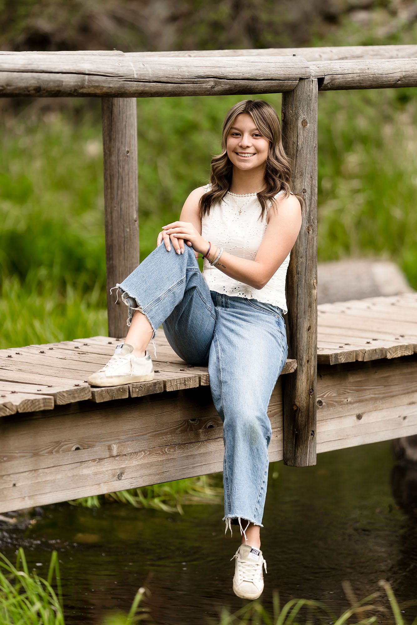 Smiling teen girl in jeans and white top sitting on rustic wooden bridge over stream.