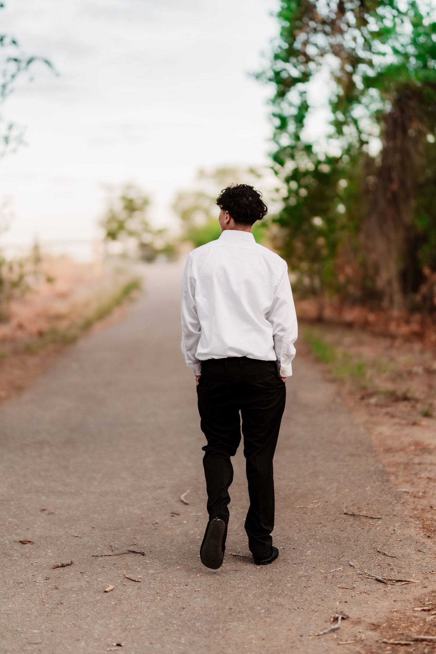 Person in white shirt and black pants walking alone on a dirt path surrounded by trees at sunset.