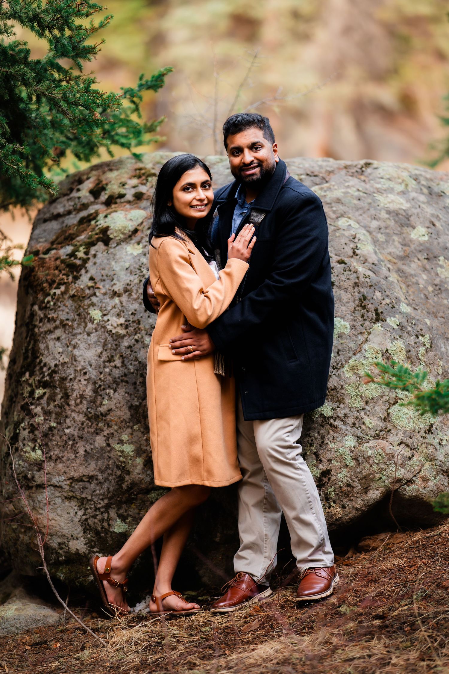 Couple in elegant winter attire pose together against large boulder in natural forest setting.