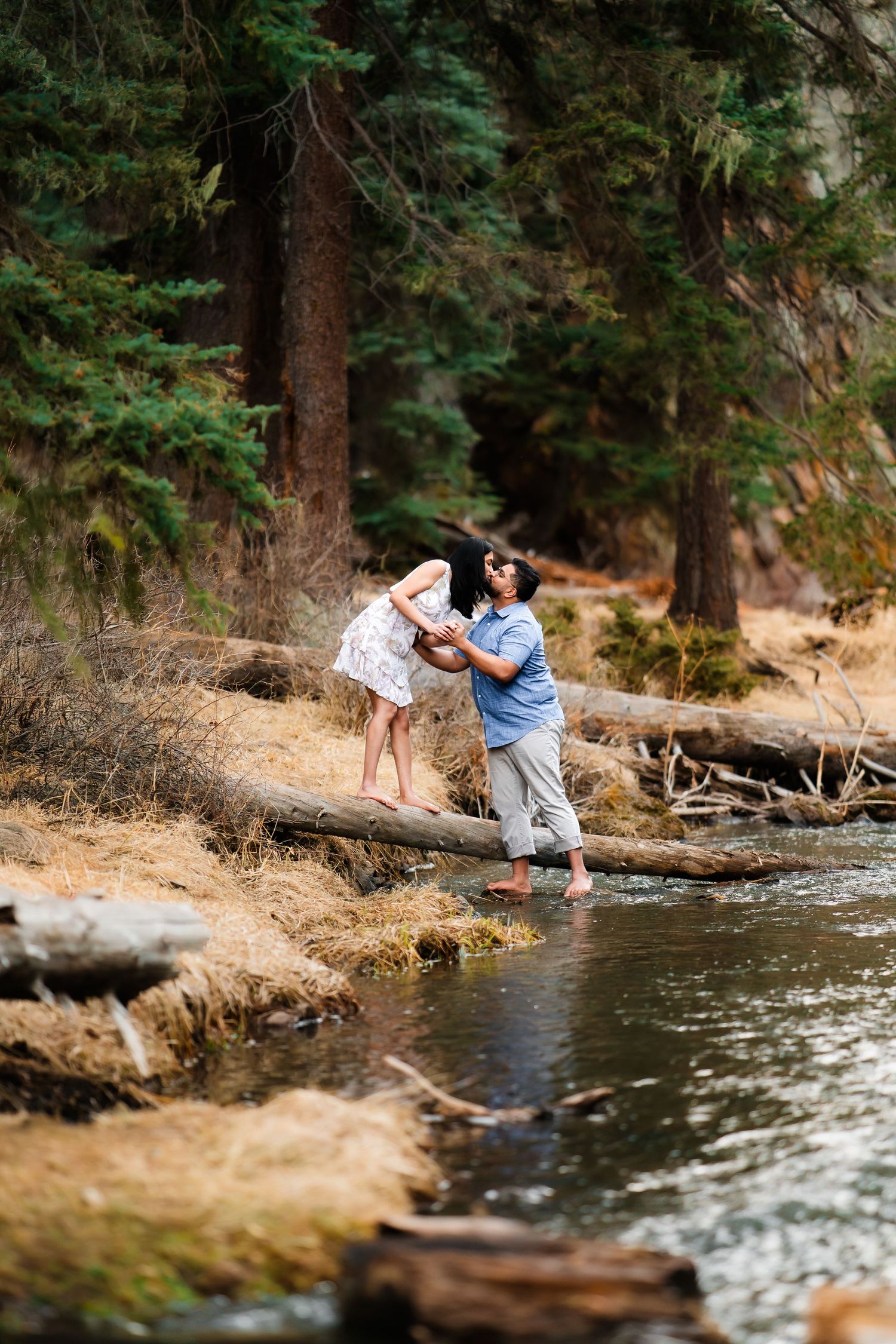 A couple balances carefully on fallen logs while crossing a stream in a forested area.