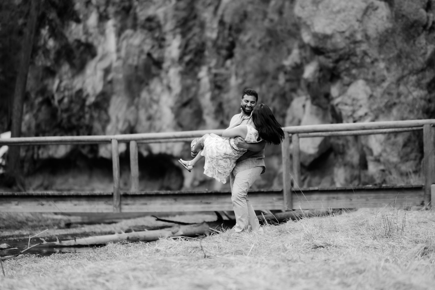Black and white romantic photo of a couple sitting together on a wooden railing in a natural outdoor setting.