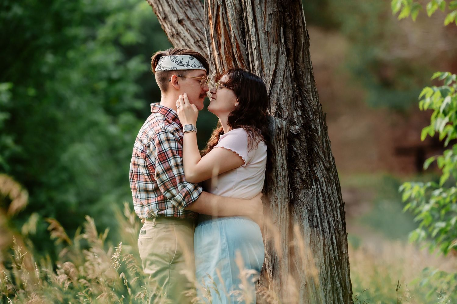 A romantic couple embraces near a rustic tree trunk in a natural outdoor setting with soft lighting.