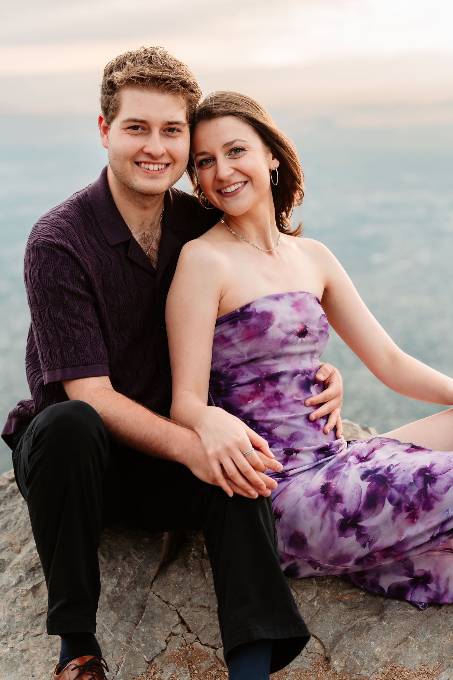 A couple sits together on rocky terrain wearing purple and black attire during an outdoor photoshoot.