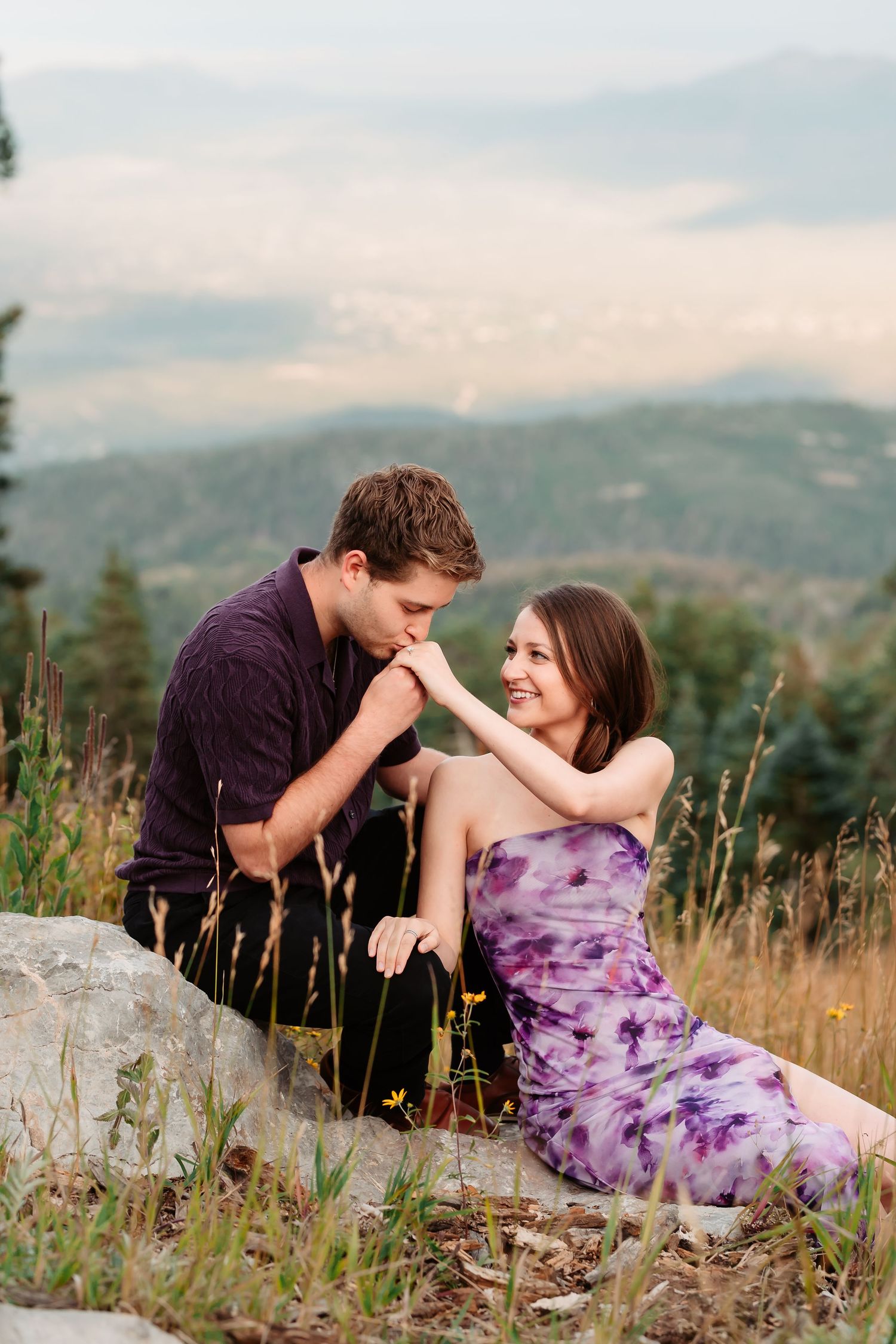 A romantic couple sits on a rocky outcrop surrounded by wildflowers with mountains in the background.