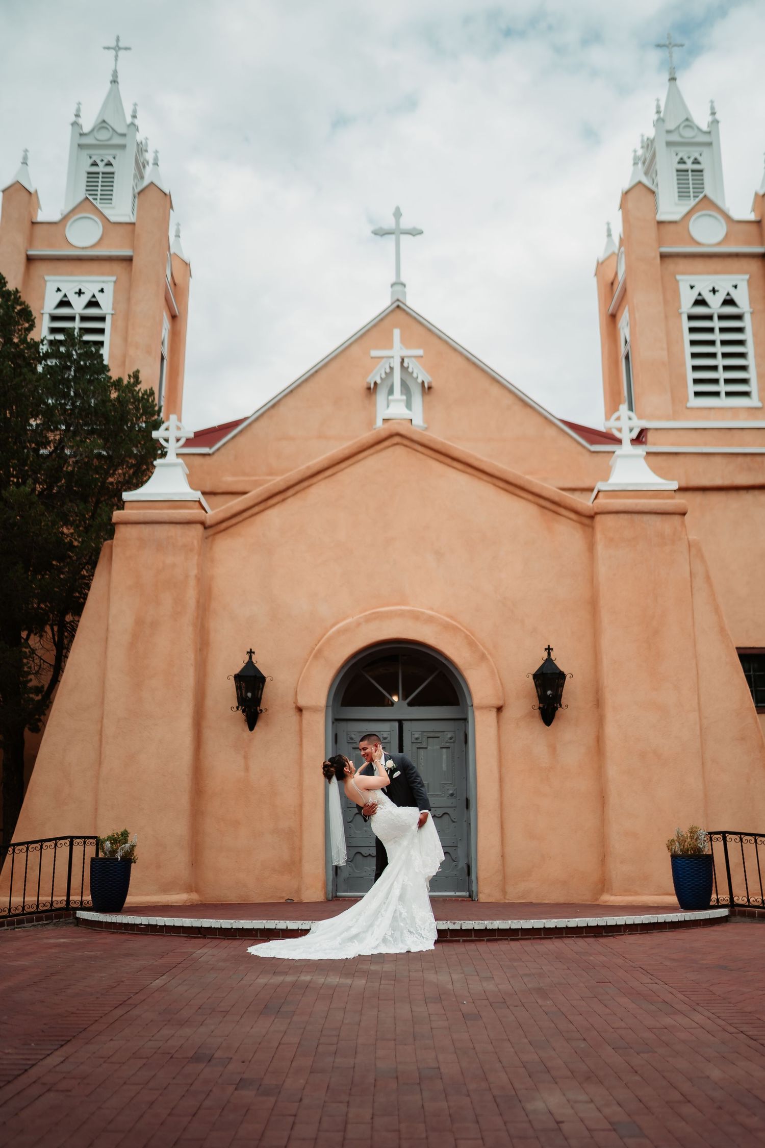 Wedding couple shares romantic moments in front of a historic adobe church with twin bell towers and arched entrance.