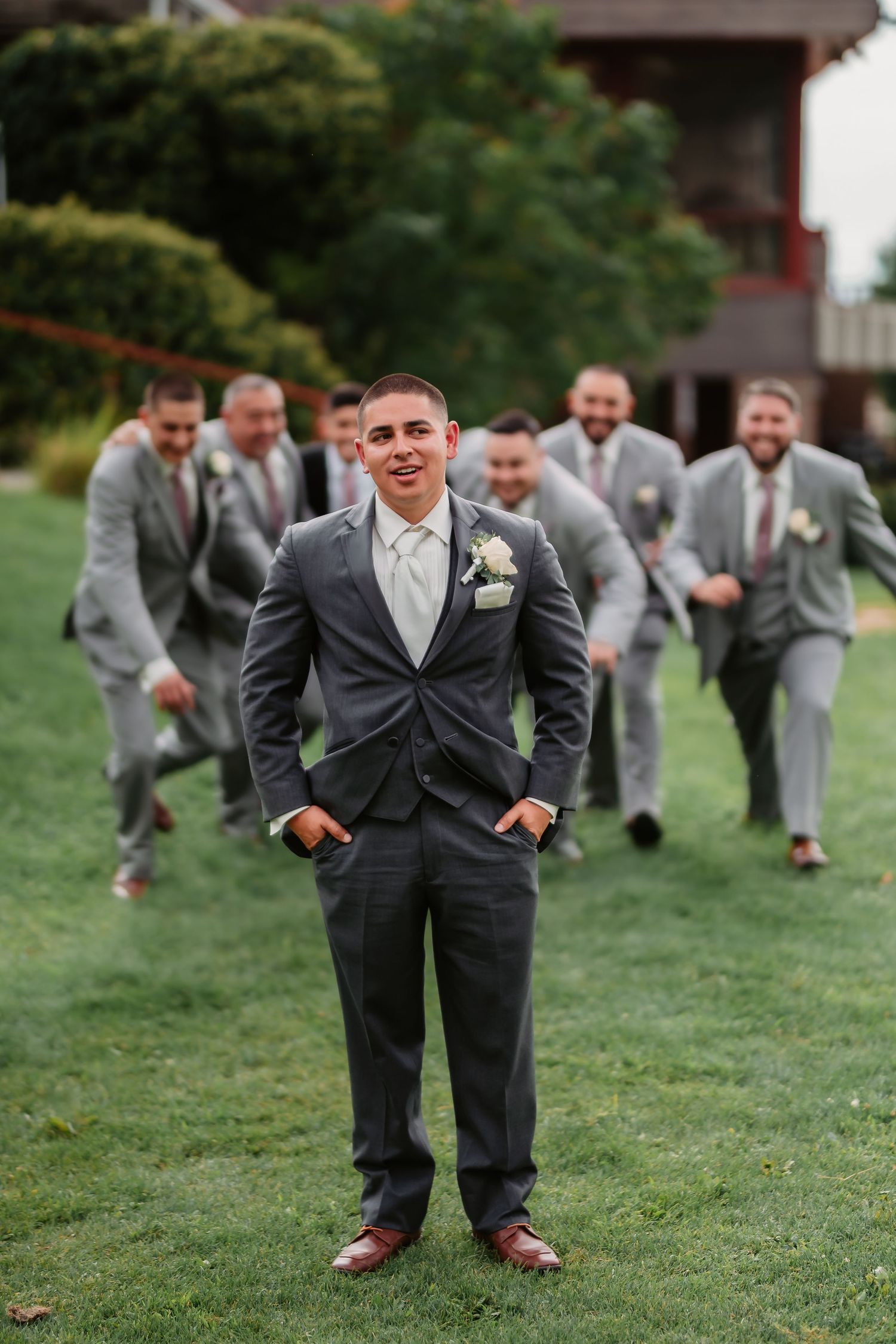 A playful wedding party lineup poses on grass wearing matching gray suits showing different stances.