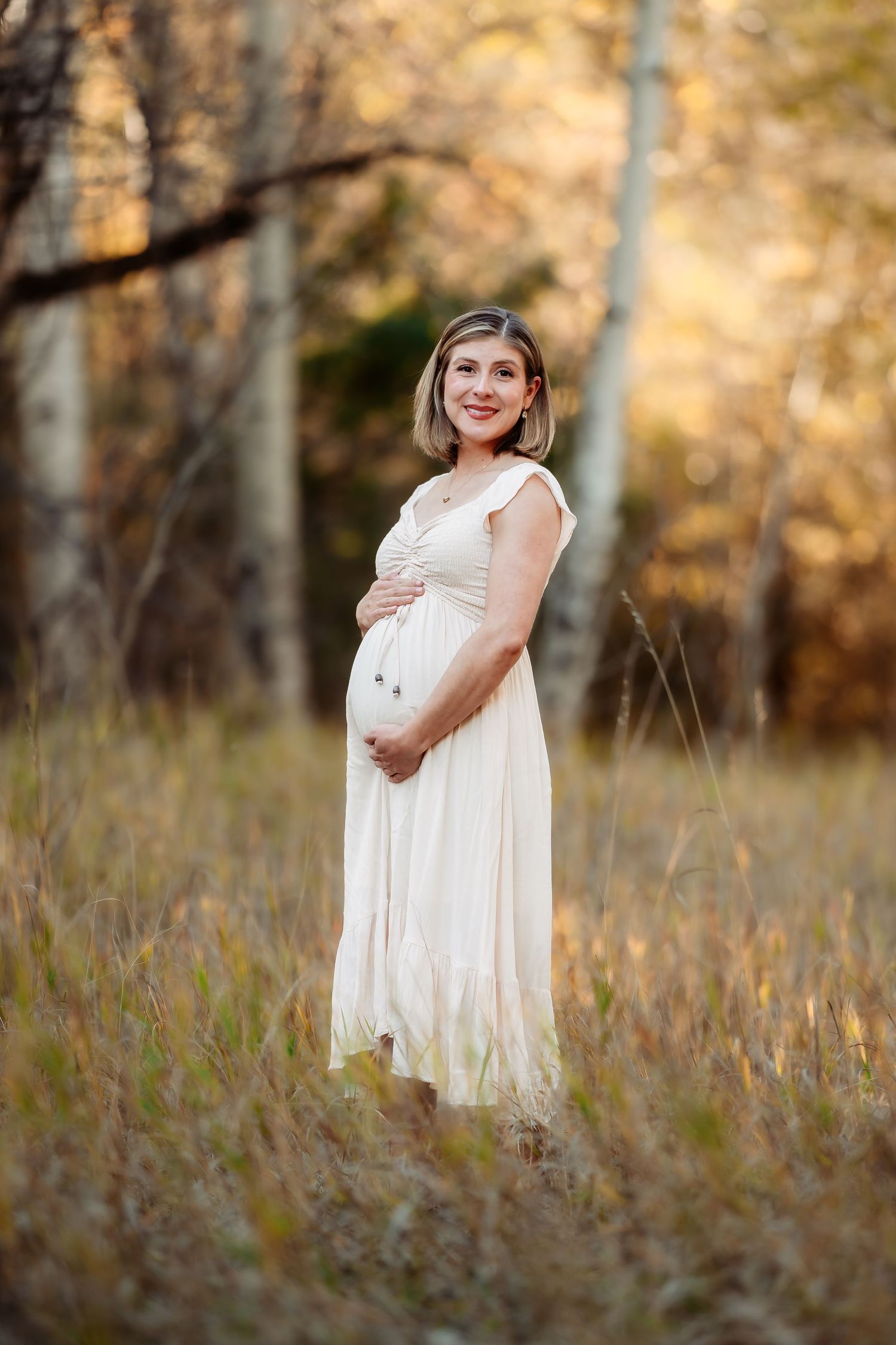 A maternity photo in a white flowing dress standing in tall autumn grass with golden bokeh background.