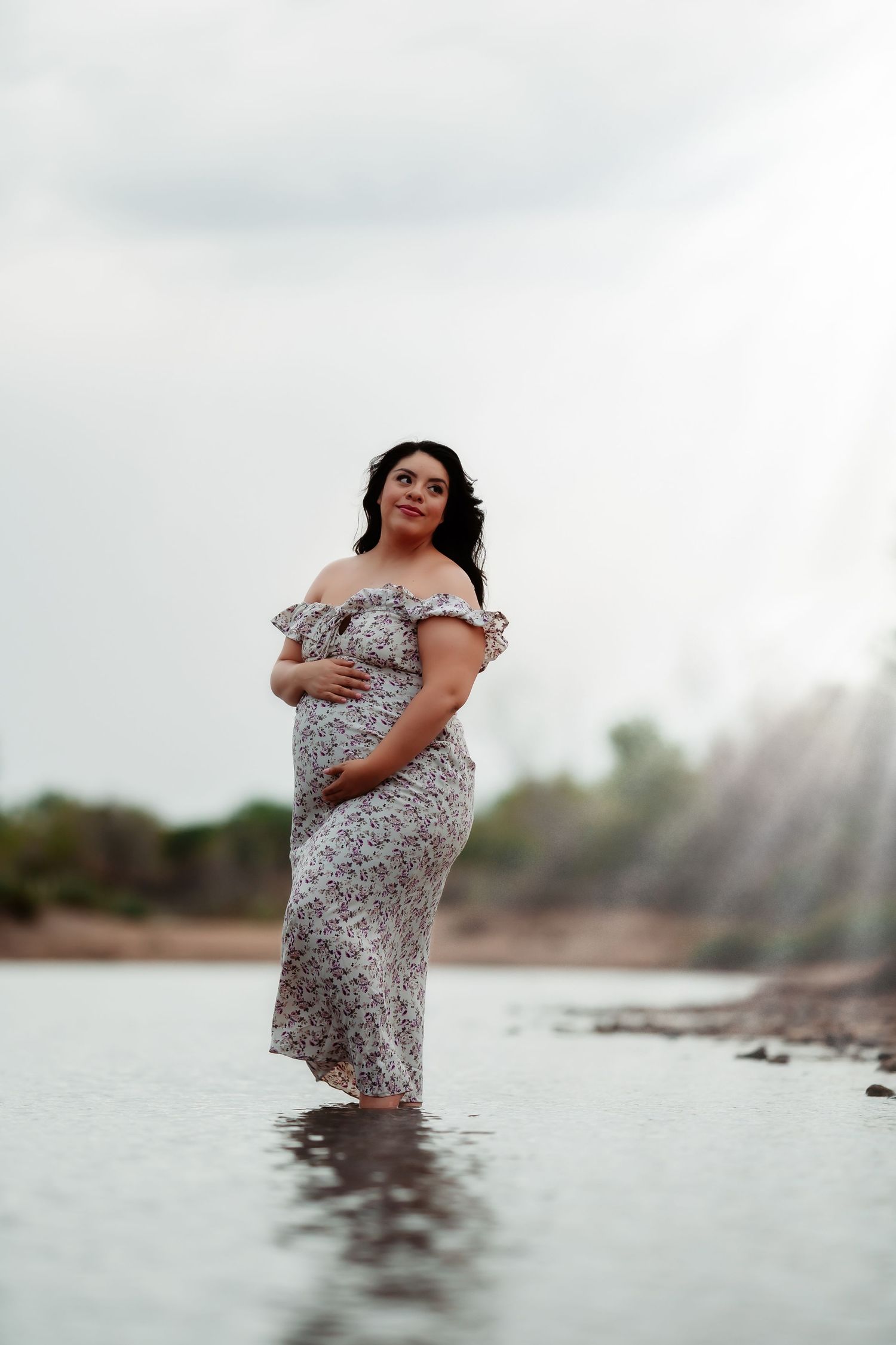 A woman in a floral dress stands in shallow ocean water during a maternity photoshoot at sunset.