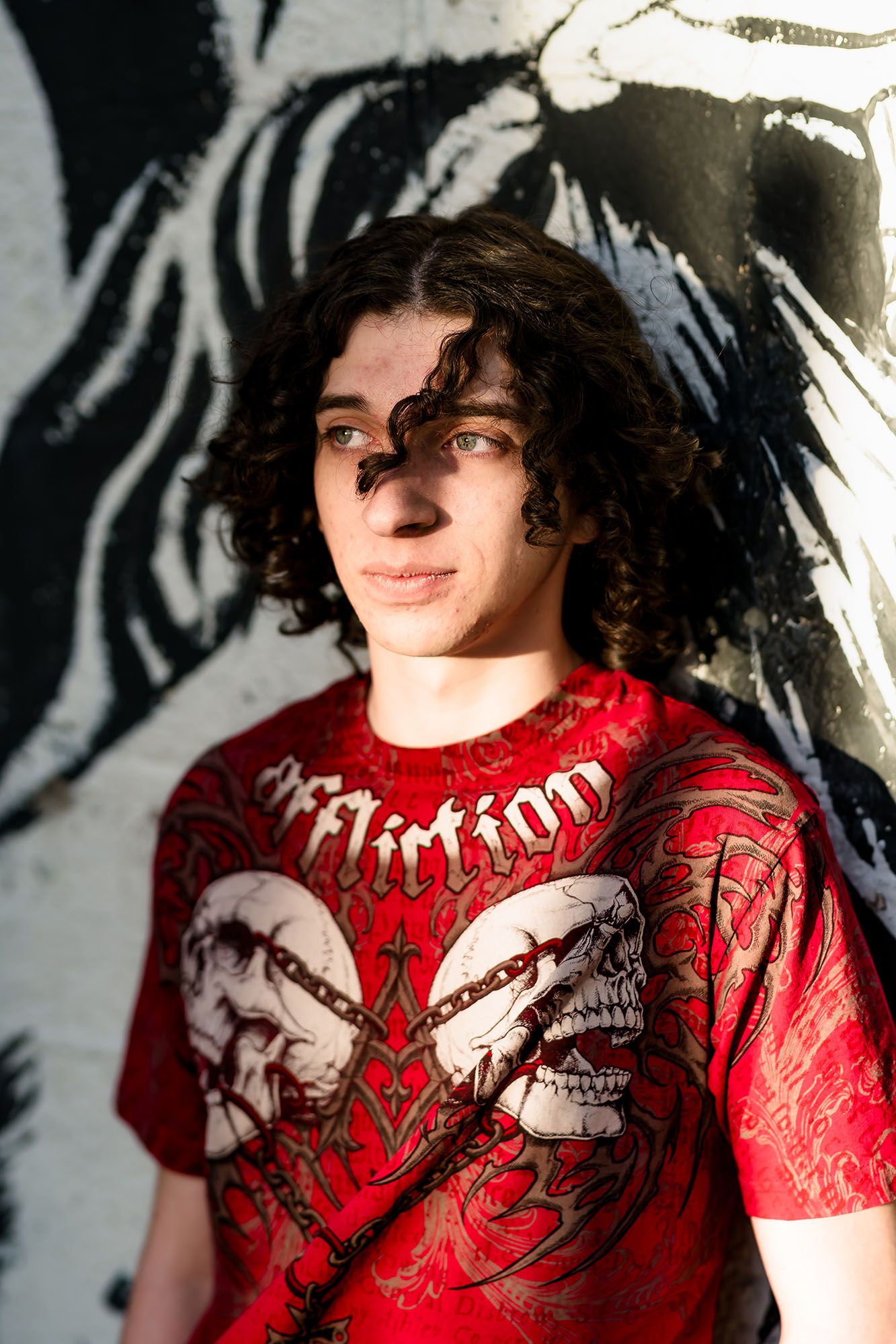 Young man with curly hair wearing red Affliction t-shirt poses against graffiti wall in sunlight.